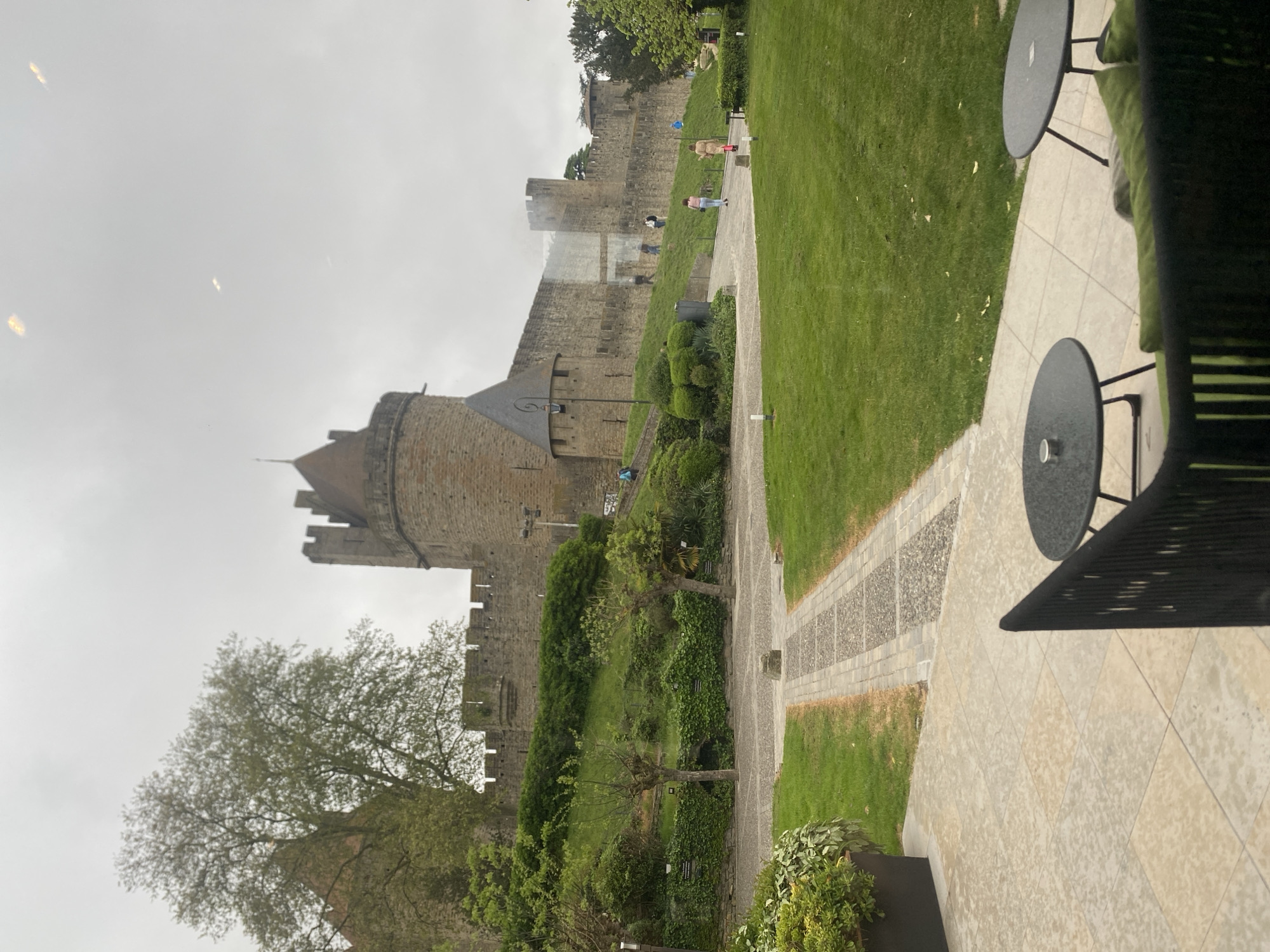 A medium, eye-level shot captures a view of Carcassonne, France, under an overcast sky. The medieval fortress's stone walls and towers dominate the left half, while a grassy slope and a stone-paved path occupy the right. The path winds alongside the fortress, with a few people walking along it. Foreground elements include outdoor seating with dark tables and chairs, partially visible on the paved patio, suggesting a café or restaurant setting. Vegetation includes trees, bushes, and grass. The overall mood is subdued due to the grey, overcast weather.