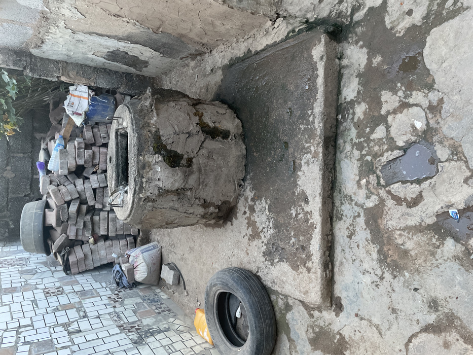 The image displays a cluttered and somewhat neglected outdoor or semi-enclosed utility area, possibly a backyard corner, storage space, or alleyway. The scene is illuminated by natural light, suggesting daytime, without harsh shadows.
In the center of the frame, a large, rough concrete cylinder or pipe segment lies horizontally on the ground. It appears old and weathered, with dark stains and patches of moss or grime clinging to its surface. One end of the cylinder is visible, revealing a hollow interior and what looks like some protruding metal rods or rebar.
To the left of the concrete cylinder, there is a large, irregularly stacked pile of light grey, interlocking pavers or bricks. Some plastic bottles and small pieces of debris are mixed in with the bricks. Behind this pile, a black tire or wheel rim is partially visible, standing upright against a concrete or block wall. Further left and towards the bottom, the ground transitions to an area paved with white and grey square tiles. A white sack, possibly containing cement or other building material, is situated near the base of the brick pile, with the word "BANCUT" clearly visible on its side. Some green foliage from a plant or bush is visible in the upper left corner, growing near the wall.
To the right of the concrete cylinder, a flat, rectangular concrete slab or raised platform extends from a wall. This slab, along with the surrounding ground, is discolored and stained, with several puddles of water indicating dampness. On the lower right side of the foreground, another black tire lies flat on the ground next to a bright yellow plastic jerry can. Scattered across the concrete ground and within the puddles are various small pieces of trash and debris, including wrappers and plastic fragments.
The walls in the background and to the right are made of rough concrete or plastered material, showing signs of wear, cracking, and peeling. An overhead concrete structure or ceiling is also visible at the top of the image, similarly aged and discolored. The overall impression is one of disuse, wear, and a lack of maintenance.