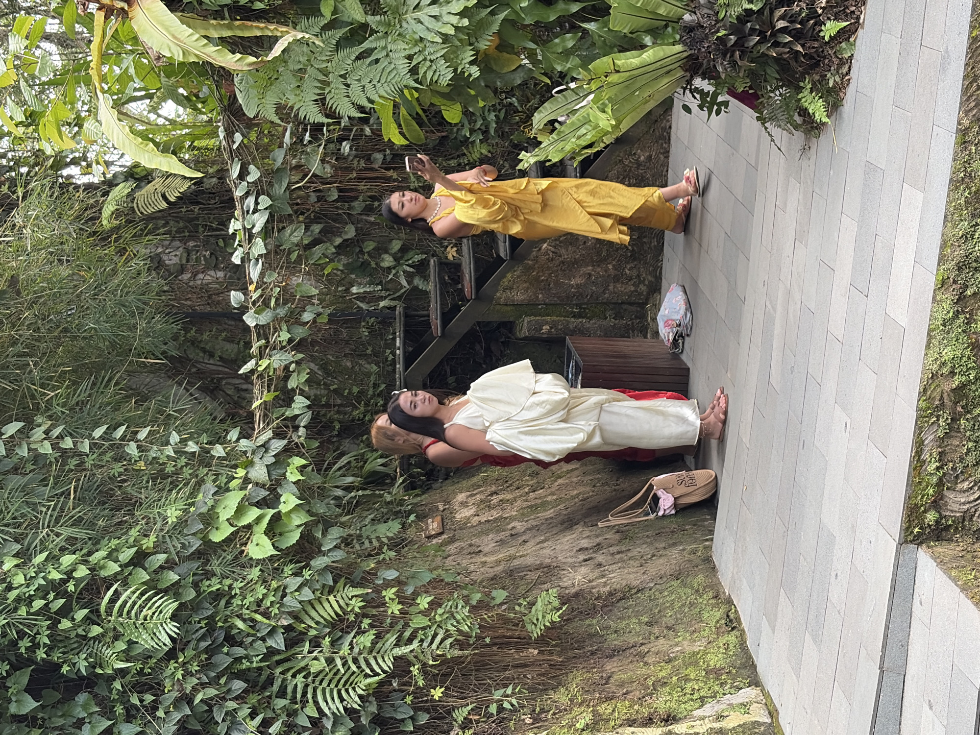 The image depicts a scene in the Tegallalang Rice Terraces in Tegallalang, Indonesia. Three individuals are present. On the left, two women stand near a stone wall covered in lush greenery and hanging vines. One woman, with blonde hair, wears a red dress, while the woman in front of her, with dark hair, is dressed in a flowing white garment and holds a white cloth. A woven bag with a pink accessory hangs from the wall behind them. To the right, a woman with dark hair, wearing a bright yellow outfit and a pearl necklace, is holding up a smartphone to take a picture or video. She is standing on a paved walkway that runs alongside the verdant natural environment. Stairs made of stone and wood are visible behind the women, leading upwards into the dense foliage. The overall atmosphere is one of tropical beauty and leisurely exploration.