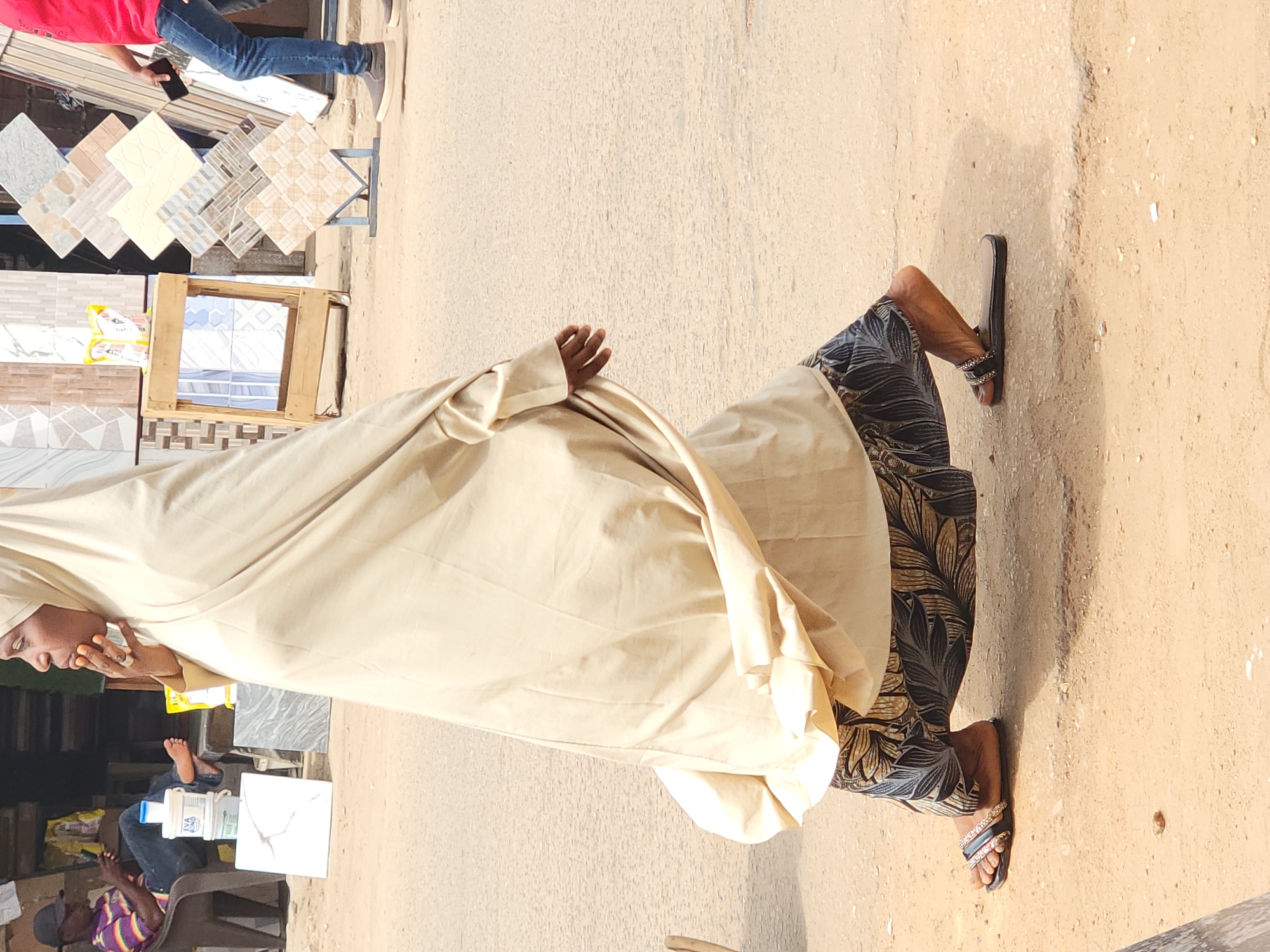 The scene is an outdoor street in Maiburiji, Nigeria. A woman in a light cream-colored hijab and patterned pants stands on a dusty sidewalk. She is wearing sandals. Nearby, several tiles are displayed on a stand, some with varying patterns and colors. Other people are visible in the background, including someone wearing a red shirt and jeans and someone sitting on a chair wearing a striped shirt. There are various objects such as tiles, wooden crates, and plastic containers. The overall setting suggests a marketplace or roadside shop.