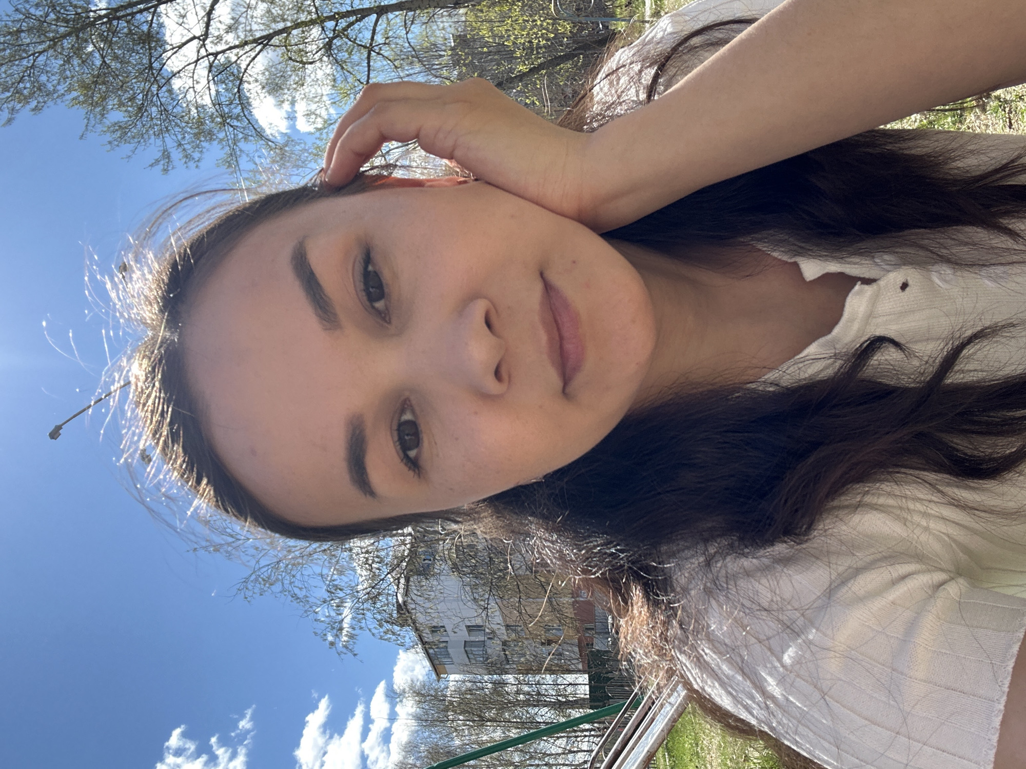 A woman is taking a selfie outdoors. The background consists of a blue sky with scattered clouds, tree branches, a building with windows, and green vegetation. Her hair is dark. She is wearing a white top. Her hand is near her ear. The location is Uray, Russia.