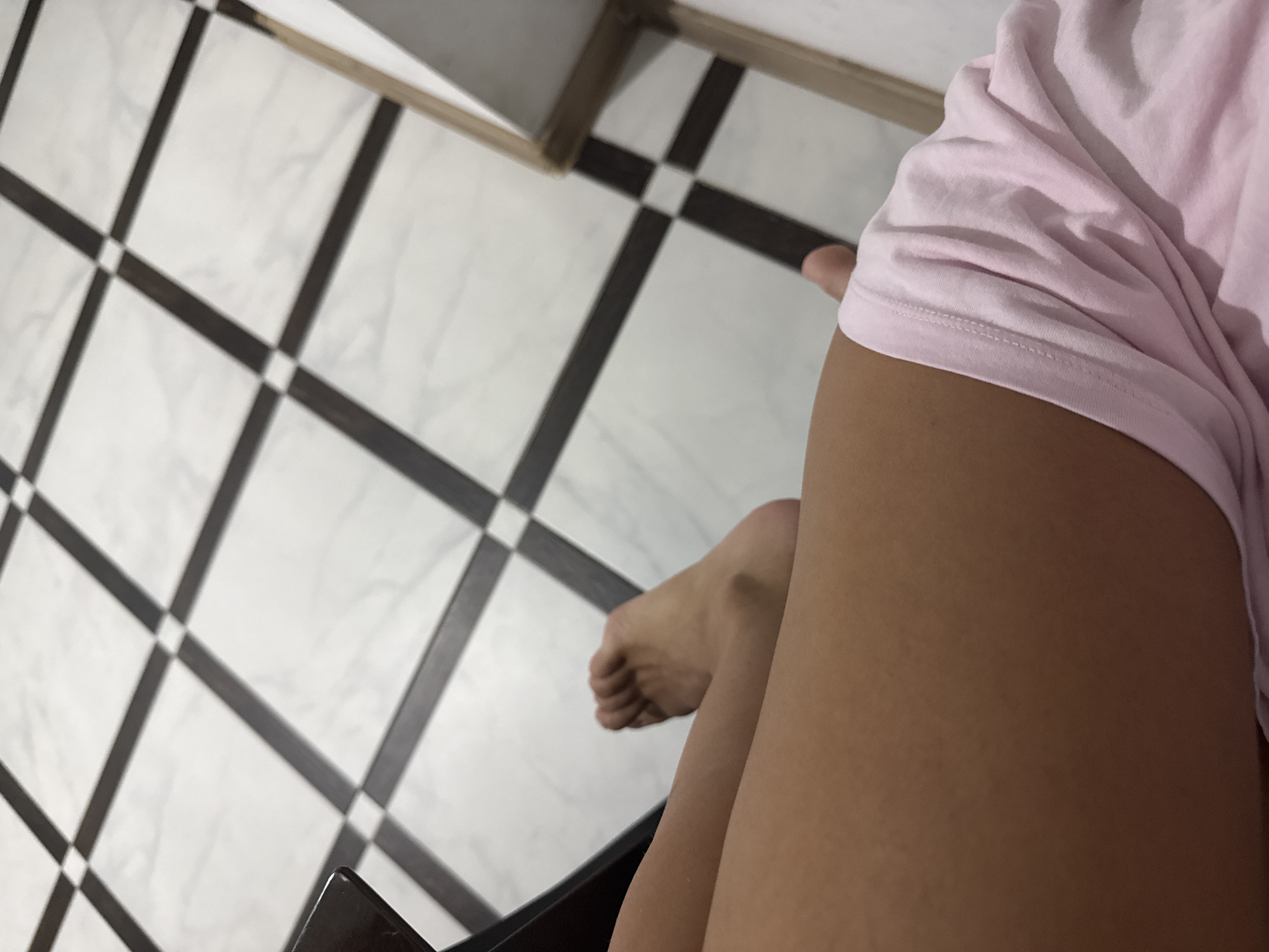 The image shows a close-up view of a person's legs and feet. The person is sitting, with their right leg bent at the knee and their foot resting on the floor. They are wearing a light pink garment, possibly shorts or a skirt. The floor is tiled with a pattern of white and dark squares. A dark object, possibly a table or chair, is visible in the upper left corner. The scene is indoors, and there is no indication of the specific location within Chișinău, Moldova.