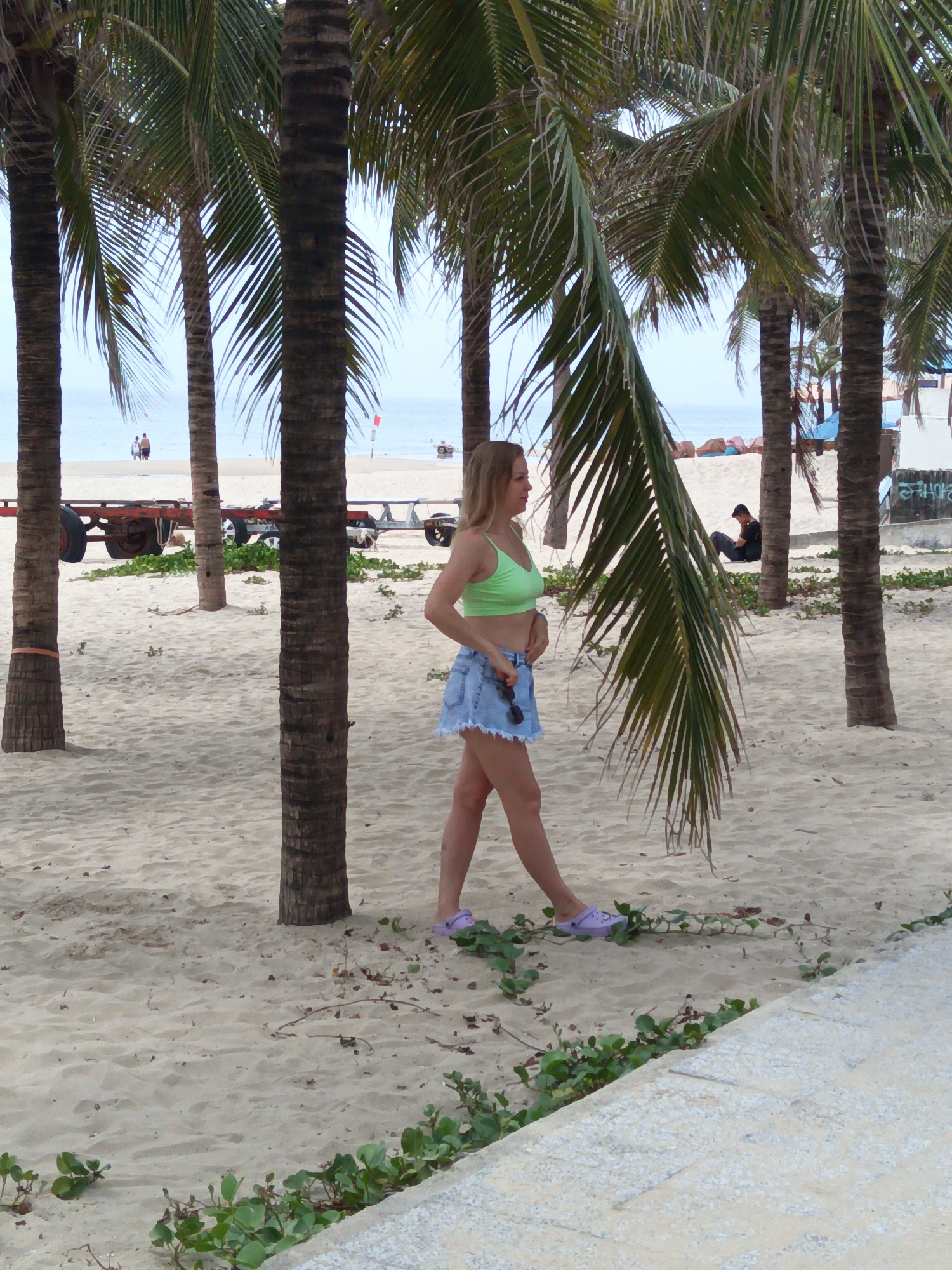 In Đà Nẵng, Vietnam, on a sandy beach lined with palm trees, a young woman stands facing left. She wears a light green crop top and a short denim skirt, holding a pair of sunglasses. Her feet are clad in light purple clogs. In the background, the ocean stretches to the horizon under a pale sky. Further back, several palm trees stand, some with orange banding on their trunks. A trailer with large wheels is parked on the sand. Two people are visible in the distance near the water, and another person sits on the sand beneath a palm tree. Green vines grow along the edge of a paved walkway in the foreground.