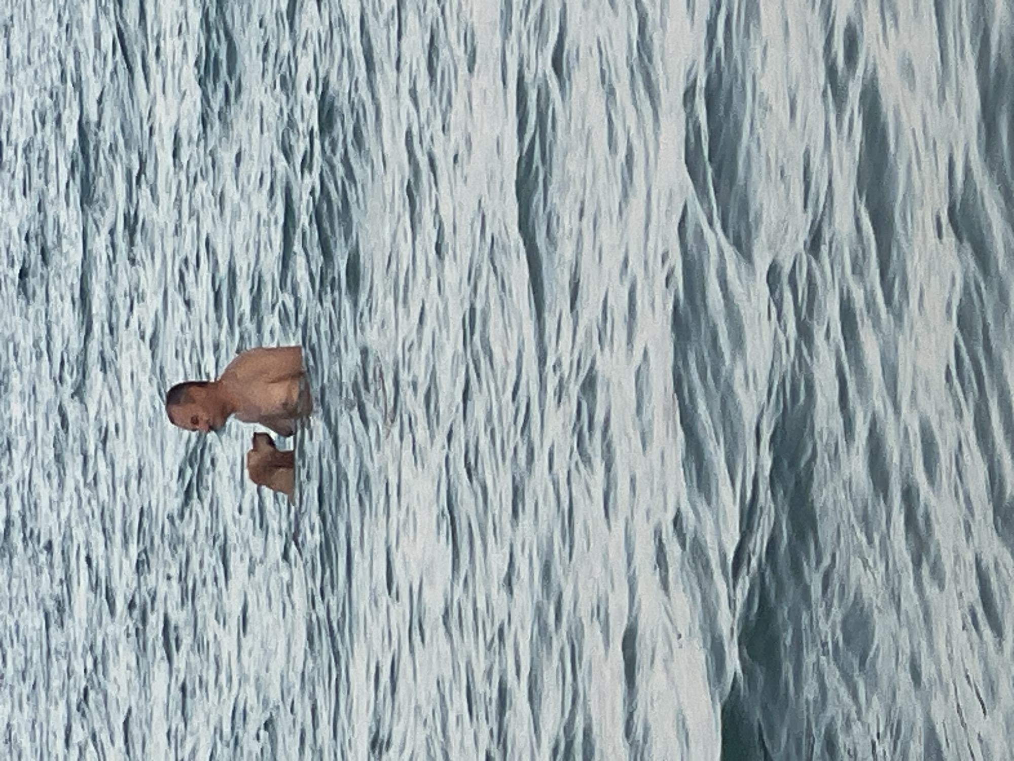 An overhead, medium shot captures a middle-aged, shirtless man and a tan dog in the ocean. The man, facing right, stands in the water, his chest and head visible above the surface. The dog, positioned to his left, is also partially submerged, with its head turned towards the man, suggesting interaction. The water is a muted blue-gray, with subtle waves creating a textured surface. The background is entirely composed of this rippling water, with no land or sky visible. The image appears to have been taken in the ocean waters off the coast of Trikomo, Northern Cyprus.