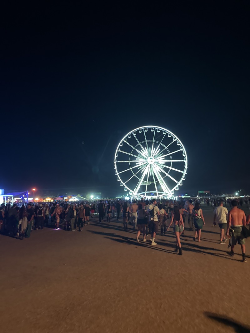 The image captures a night scene at the Coachella Valley Music and Arts Festival in Indio, California, United States. A large Ferris wheel, illuminated with bright white lights, dominates the background. A dense crowd of people is visible in the foreground and middle ground, spread across a sandy or dirt-covered area. The crowd's attire varies, suggesting casual festival wear. In the distance, structures and lighting indicate the presence of stage areas or vendor booths. The sky is dark.