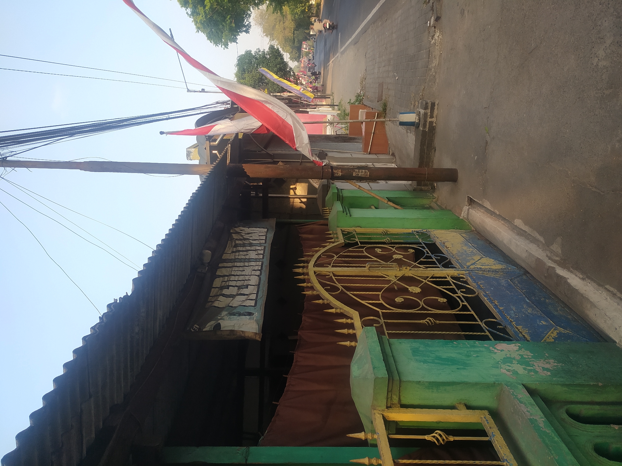 The image depicts a street scene in Tanggul, Indonesia, during daylight. In the foreground, a green and yellow painted gate with intricate metalwork is visible, with a blue section at its base. Behind the gate, a building with a corrugated metal roof and brown curtains is present. A banner with text, partially obscured, hangs above the curtains. Two Indonesian flags, red and white, are prominently displayed on flagpoles.
The street itself is paved with interlocking bricks and asphalt. A utility pole with numerous wires runs vertically along the right side of the image. Several people are seen in the distance, some riding motorcycles on the road. Trees line the far side of the street. The sky is clear and pale blue, indicating fair weather. The overall scene suggests a typical urban environment in Indonesia, possibly during a period of national celebration due to the flags.