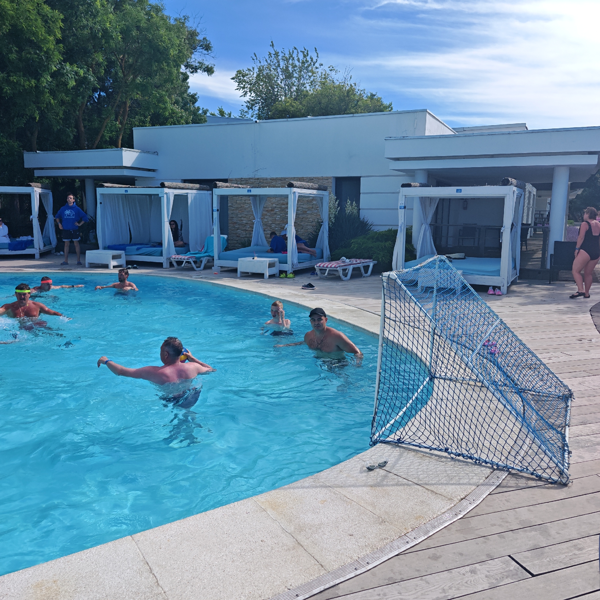 The image captures a lively scene at an outdoor swimming pool on a sunny day. The pool is filled with clear blue water, and several people are enjoying themselves. In the foreground, a man in the pool holds a yellow and blue ball, looking towards the right of the frame. To his left, another man with a yellow headband is partially submerged in the water, with ripples indicating movement. In the background, several white cabana-like structures with draped curtains line the edge of the pool, suggesting a resort or hotel setting. Some people are relaxing in these cabanas, while others are visible in the pool. A man in a blue hooded sweatshirt stands near the cabanas on the left, looking up. The overall atmosphere appears relaxed and recreational. The presence of a blue soccer goal net near the edge of the pool suggests a game or activity is underway or about to begin. The sky is blue with scattered white clouds, indicating fair weather. The lighting suggests it is daytime, likely mid-afternoon. The location context provided is "городское поселение Щелкино, Ukraine," placing the scene in a specific urban settlement in Ukraine.