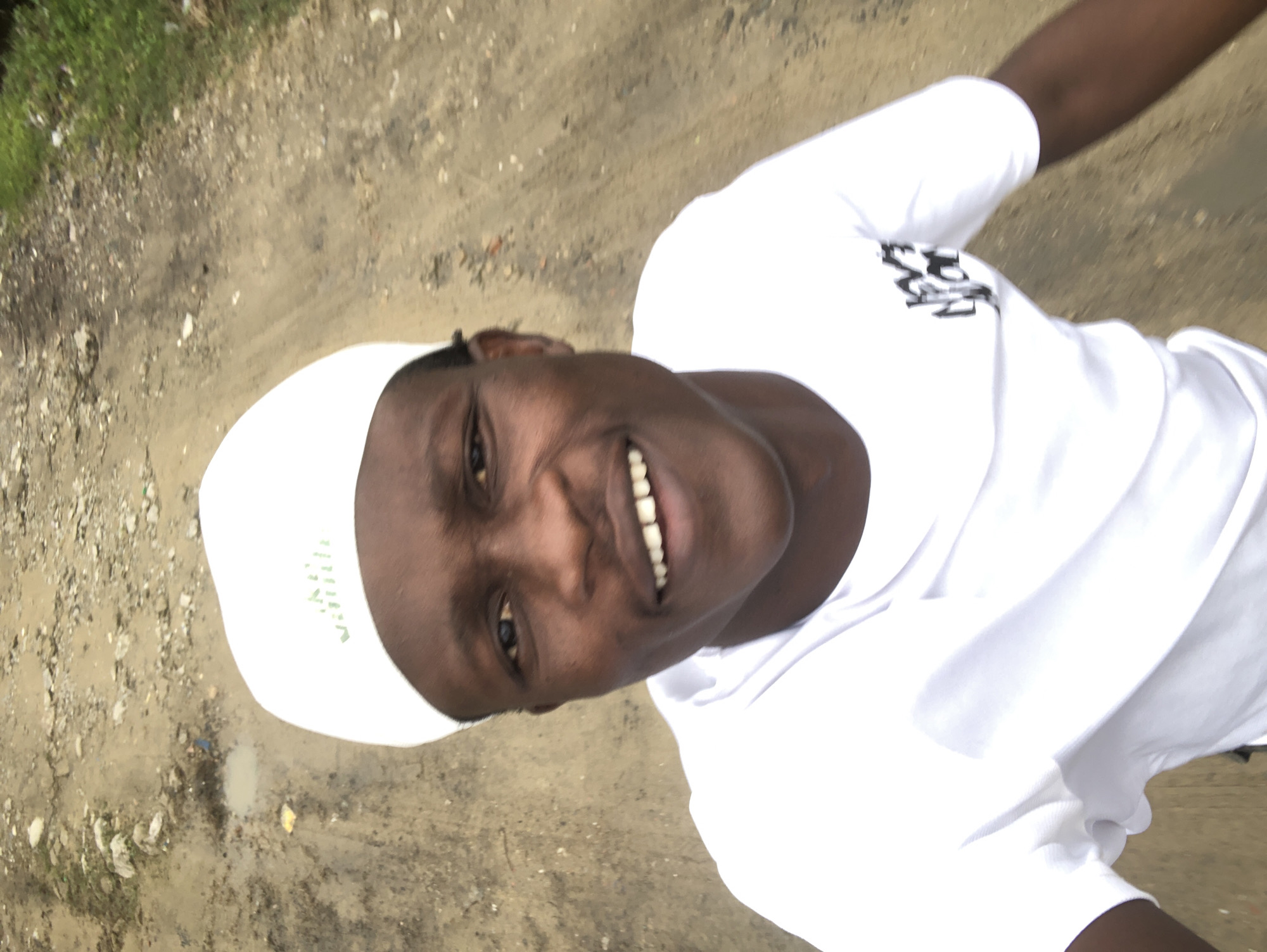 The image is a selfie taken from a high angle, showing a dark-skinned male wearing a white t-shirt and a white cap. He is smiling with his mouth open, revealing his teeth. The background is a dirt road, appearing to be a rural area with some vegetation on the right side and puddles of water on the left. The overall impression is that of a casual outdoor setting. Based on the prompt, this scene is located in Ado, Nigeria.