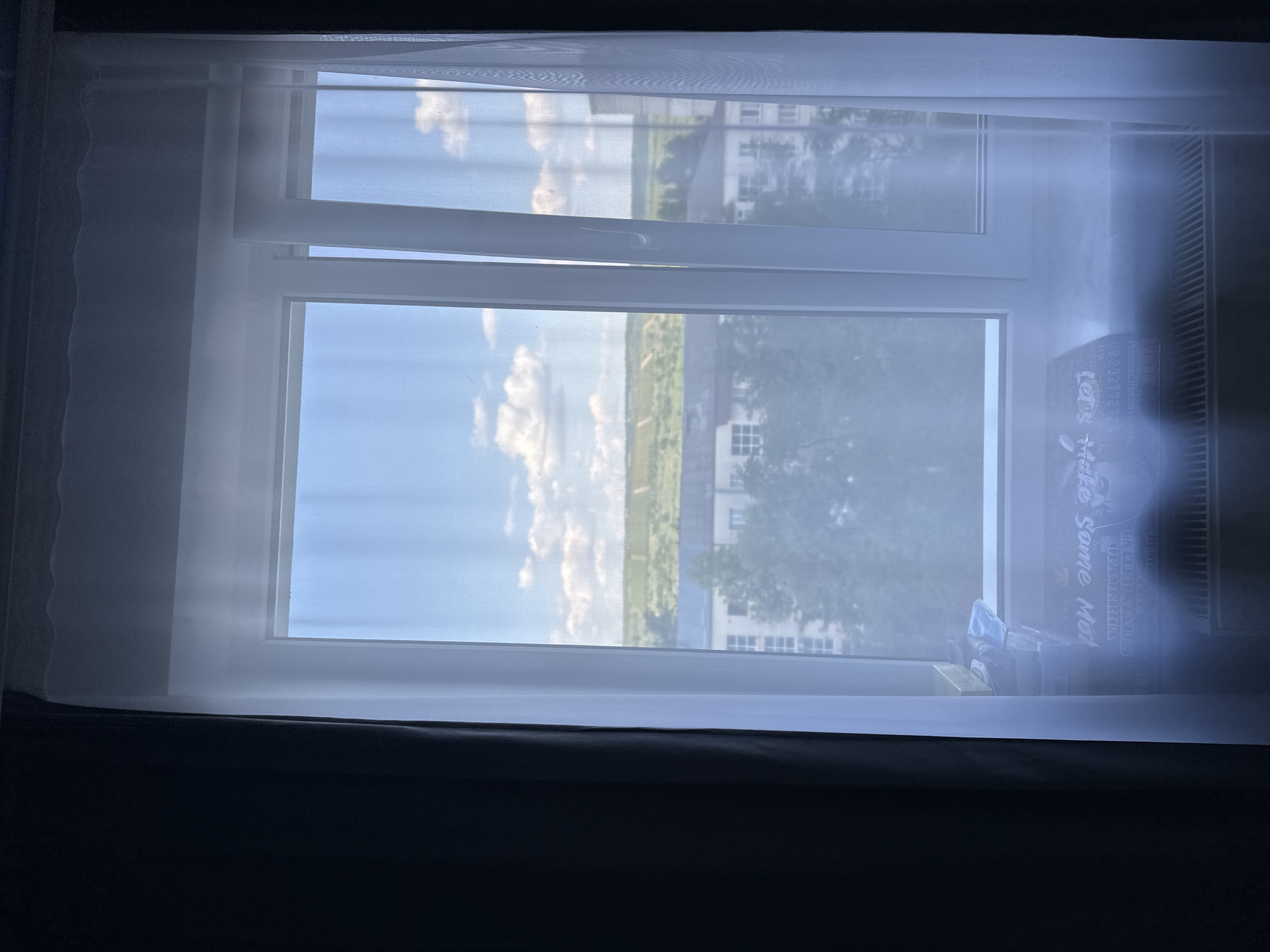 The image shows a view through a partially open window, looking out onto a bright, sunny day. The window is framed by sheer white curtains on the left and a dark curtain at the bottom. The exterior scene reveals a blue sky with scattered white clouds, a strip of green landscape, and several buildings with white facades. Trees are visible in the mid-ground, and the overall impression is one of a suburban or rural setting under a clear sky. The light suggests it is daytime, likely late morning or early afternoon. On the windowsill, near the right side of the frame, a few objects are visible, including what appears to be a box with some writing on it. The text on the box is partially obscured by glare and dust, but some words are discernible, including "Take some Mor". The setting appears to be in Strășeni, Moldova. The mood is peaceful and serene, evoked by the natural light and the quiet outdoor scene.