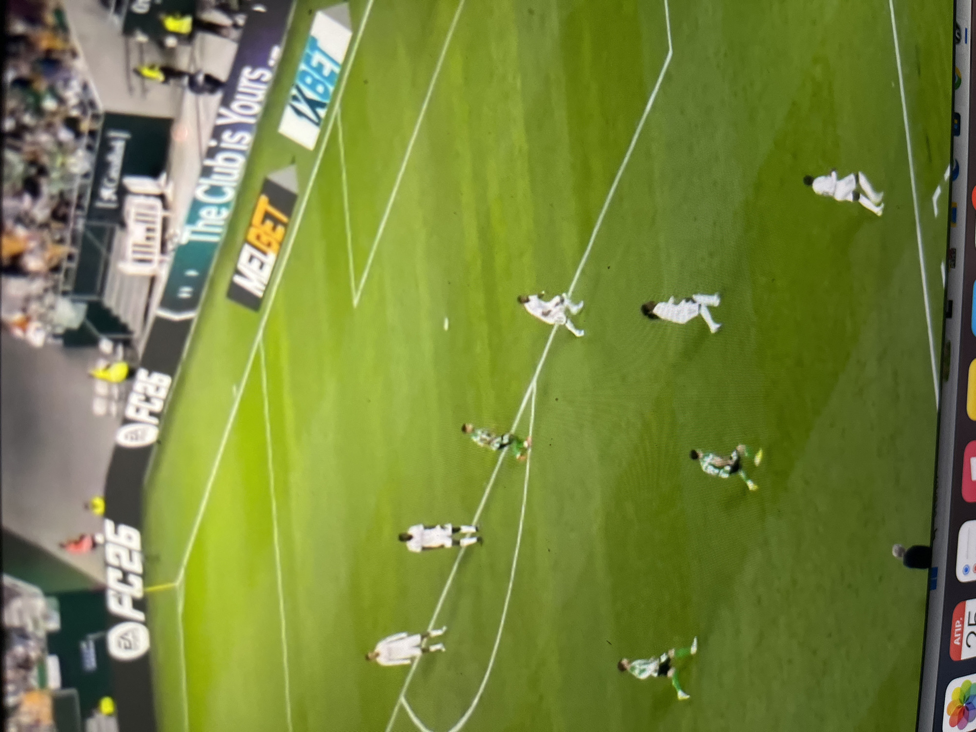 The image depicts a television screen displaying a soccer match in progress. Multiple players are visible on a green field marked with white lines, some wearing white uniforms and others green. The stadium has an advertisement banner showing the text "The Club is Yours MELBET XBET". The bottom-right corner of the screen shows an iPhone or iPad calendar application opened to April 25. There is no indication of Ulyanovsk, Russia.