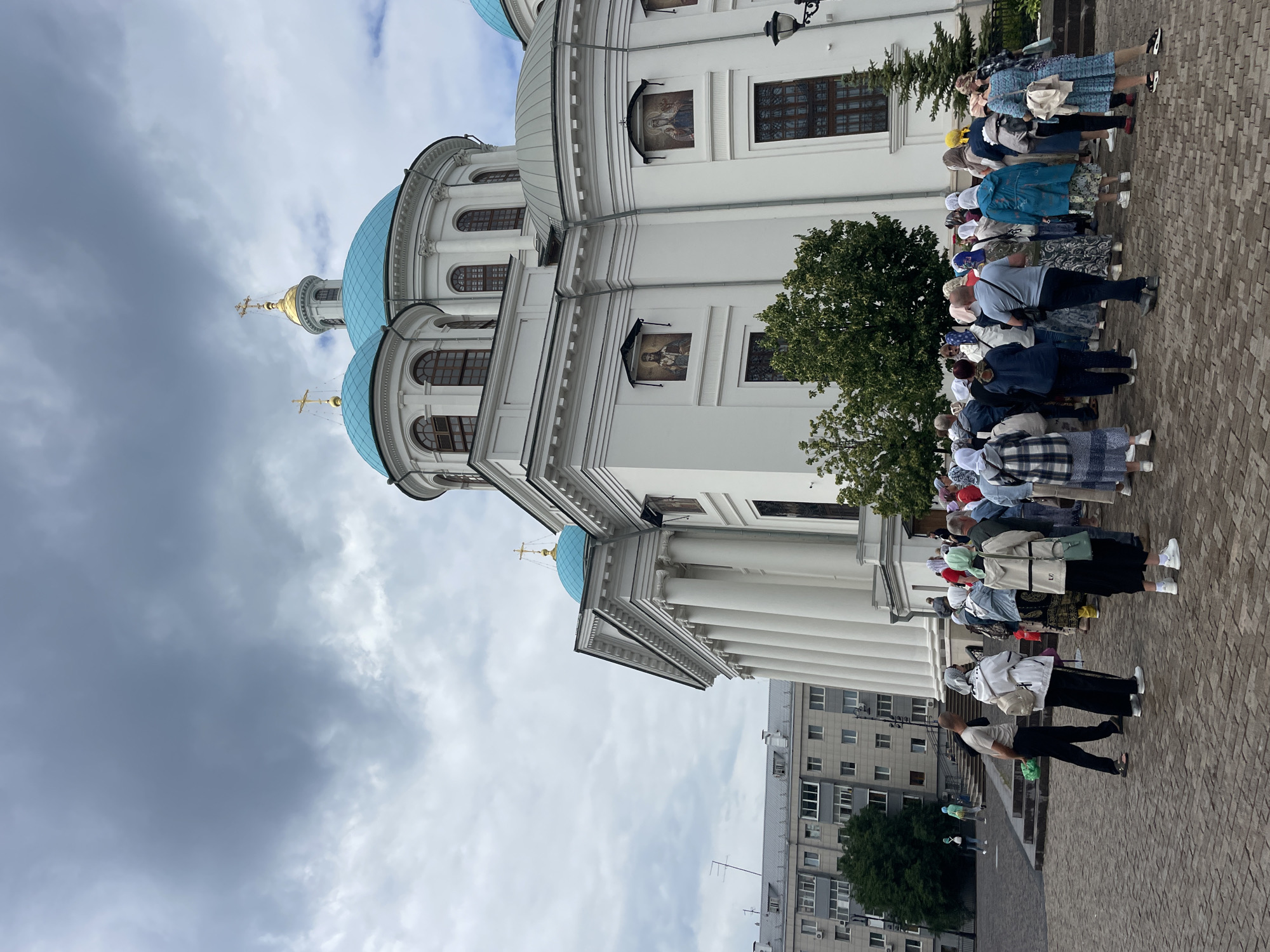 This media file captures a gathering of people outside a white church with distinctive blue domes. The church has a classical architectural style, featuring columns and arched windows. Several individuals are seen standing in a line, some appearing to be tourists or pilgrims, with many women wearing headscarves. Their postures suggest they are either waiting or observing something. The scene is set on a paved area, likely a square or walkway, with a modern apartment building visible in the background. Trees add a touch of greenery to the urban landscape. The sky is overcast with heavy, gray clouds, indicating a potentially cloudy or stormy day. The overall impression is one of a religious or cultural site visit, with the weather contributing to a somewhat subdued atmosphere. There is no visible text in the image.