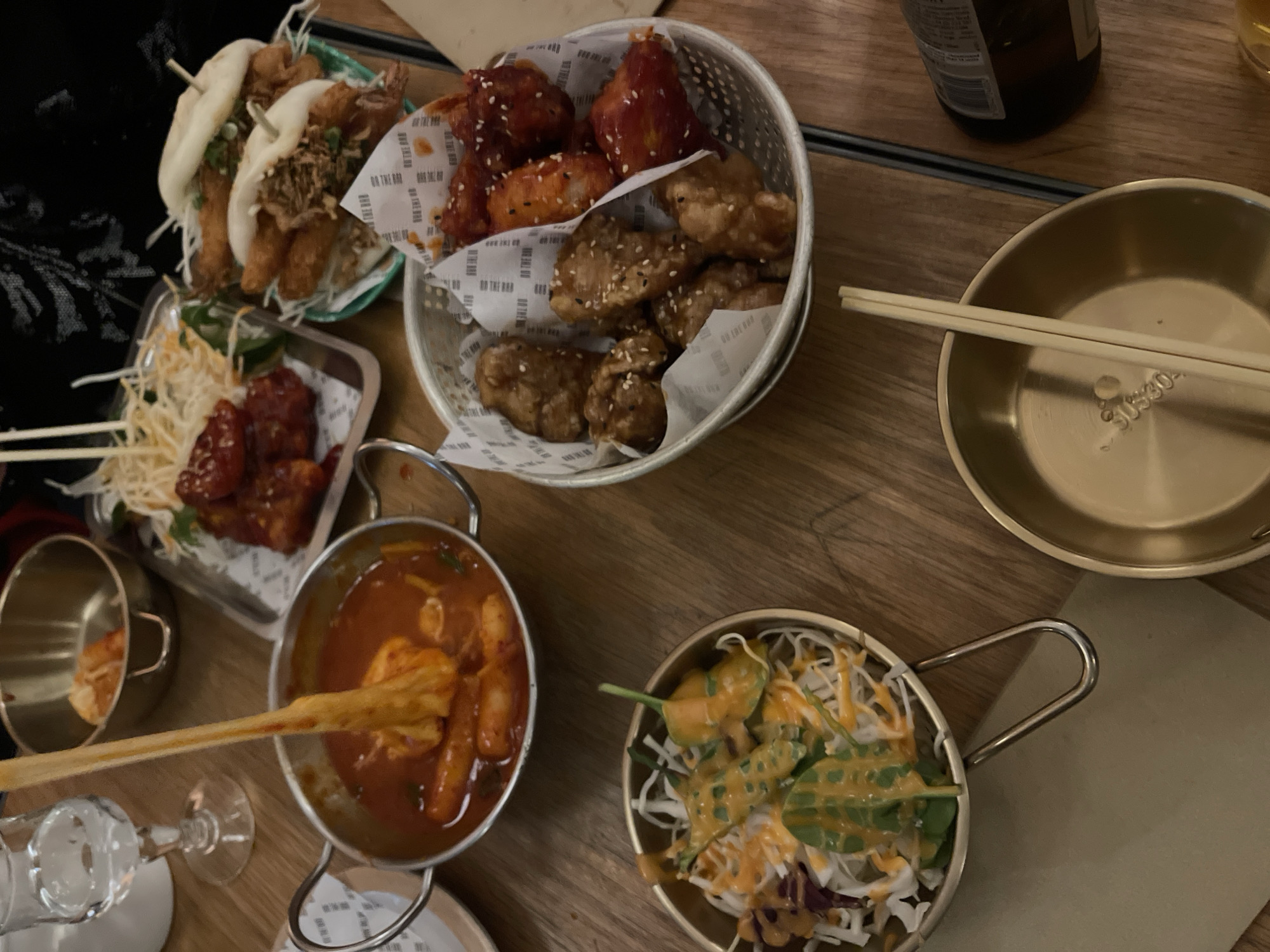 The image depicts a top-down view of a wooden table laden with various Korean dishes, possibly in a restaurant setting. There are no visible people in the image.
Food items include:
* Steamed buns with fillings, arranged on a small green plate.
* A metal basket filled with a mix of Korean fried chicken, some pieces glazed with a reddish sauce and sesame seeds, others with a darker glaze and white sesame seeds.
* A metal tray containing fried chicken with a reddish sauce and some white shredded vegetables.
* A metal pot of tteokbokki (Korean rice cakes in spicy sauce), with chopsticks partially submerged within.
* A salad in a metal pot with a creamy orange dressing.
Additionally, there's a small empty metal bowl, chopsticks, and a glass of some beverage. A dark bottle is partially visible in the upper right corner.
Given the style of the food and the apparent atmosphere, the location could potentially be in a Korean restaurant, possibly in a metropolitan area like London, United Kingdom.