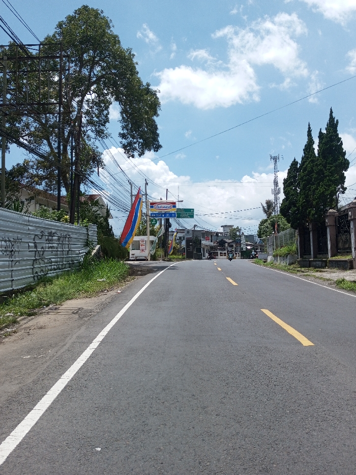 The scene depicts a daytime, wide-angle view of a two-lane asphalt road receding into the distance under a partly cloudy blue sky in Bagochen Boolingpang Ueling, Bhutan. The road features a solid white line marking the left shoulder and broken yellow lines indicating the center.
To the left of the road, a narrow dirt and grass verge runs alongside a corrugated metal fence, covered in dark graffiti. Behind the fence, a dense network of utility poles and black electrical wires is visible, intertwined with a large deciduous tree bearing green foliage. Further down the road on the left, a small white structure, possibly a vendor booth, is present, flanked by vertical red, blue, and white banners or flags. Above these, an "Indomaret" sign is mounted on a pole, and additional low-rise buildings are visible in the background.
To the right of the road, a low stone or concrete fence with decorative pillars separates the roadside from a row of tall, dark green coniferous trees. Beyond these trees, a metallic cellular tower stands against the sky. Further down the road on the right, more buildings are discernible.
A single individual riding a dark-colored motorcycle or scooter is observed in the distant center of the road, traveling away from the camera. No other persons or active vehicles are clearly visible. The overall environment suggests a semi-urban or rural thoroughfare.