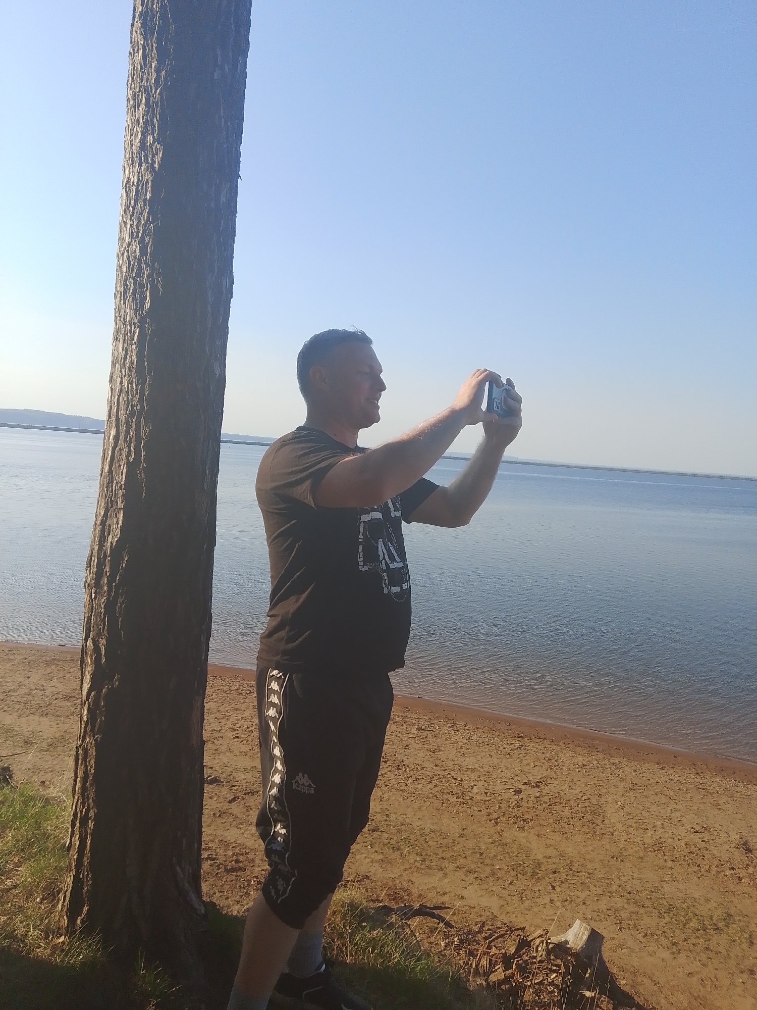 A man is taking a picture with his phone at a beach next to a large body of water. He is standing near a tree, facing the water. He is wearing a dark t-shirt and black shorts. The beach is sandy and there is a forest on the edge of the beach. The location is likely in the city of Chaikovsky, Russia, judging by the prompt.
