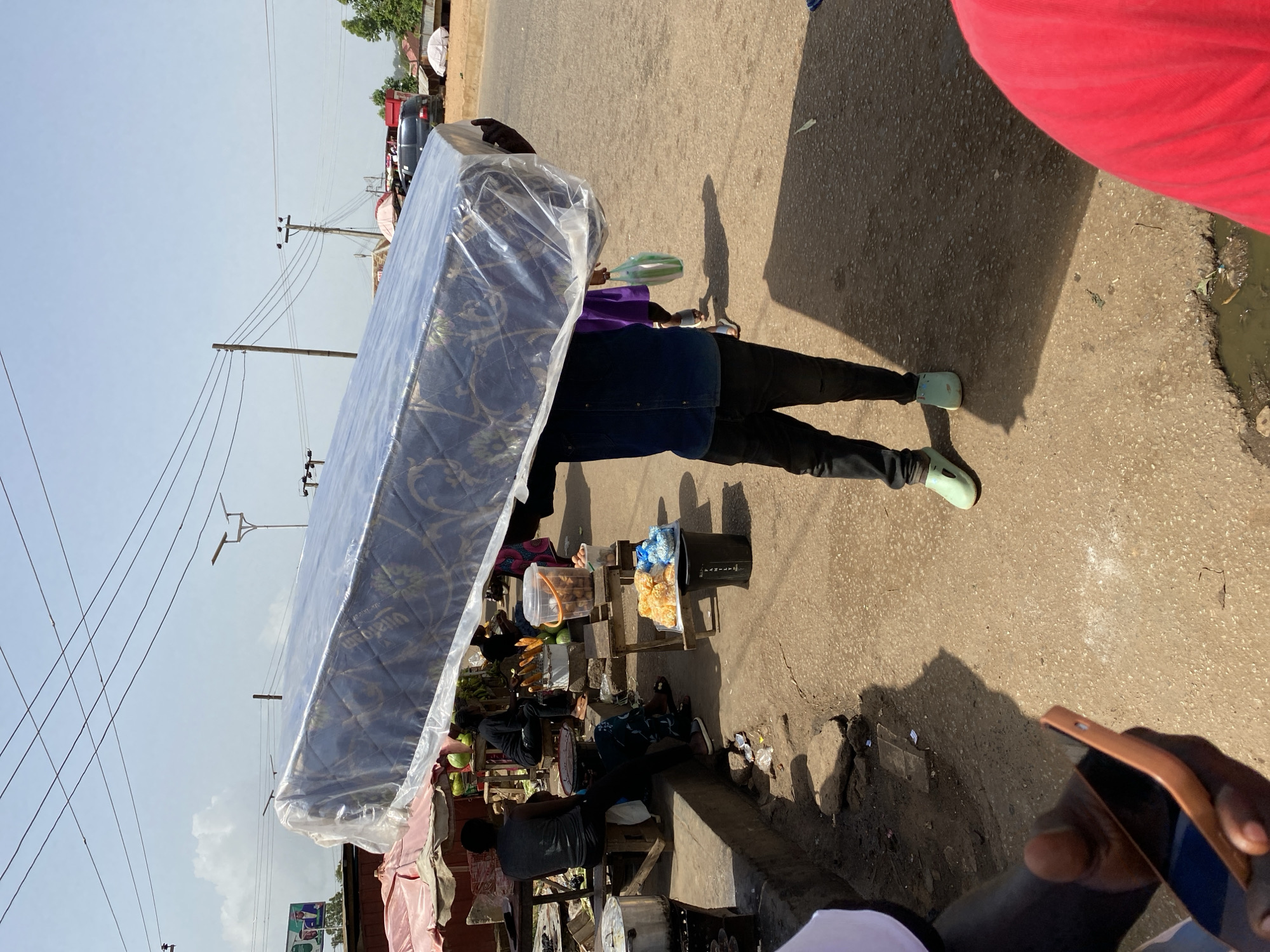 The image shows a person carrying a large mattress wrapped in plastic sheeting. The person is walking on a paved road, likely in a market setting, with a small stall of goods to their left. The stall appears to contain food items, such as fruits and what look like fried snacks. The person is wearing dark pants and light-colored shoes.
In the background, there are more market stalls, some with awnings, and a few vehicles, including a red truck. Power lines stretch across the sky, suggesting an urban or semi-urban environment. The lighting indicates it is daytime, and the presence of shadows suggests it is either morning or afternoon. The overall atmosphere seems to be that of a busy market day. A hand holding a smartphone is visible in the bottom right corner, suggesting someone is taking the photograph. The location context is Bukuru, Nigeria.
