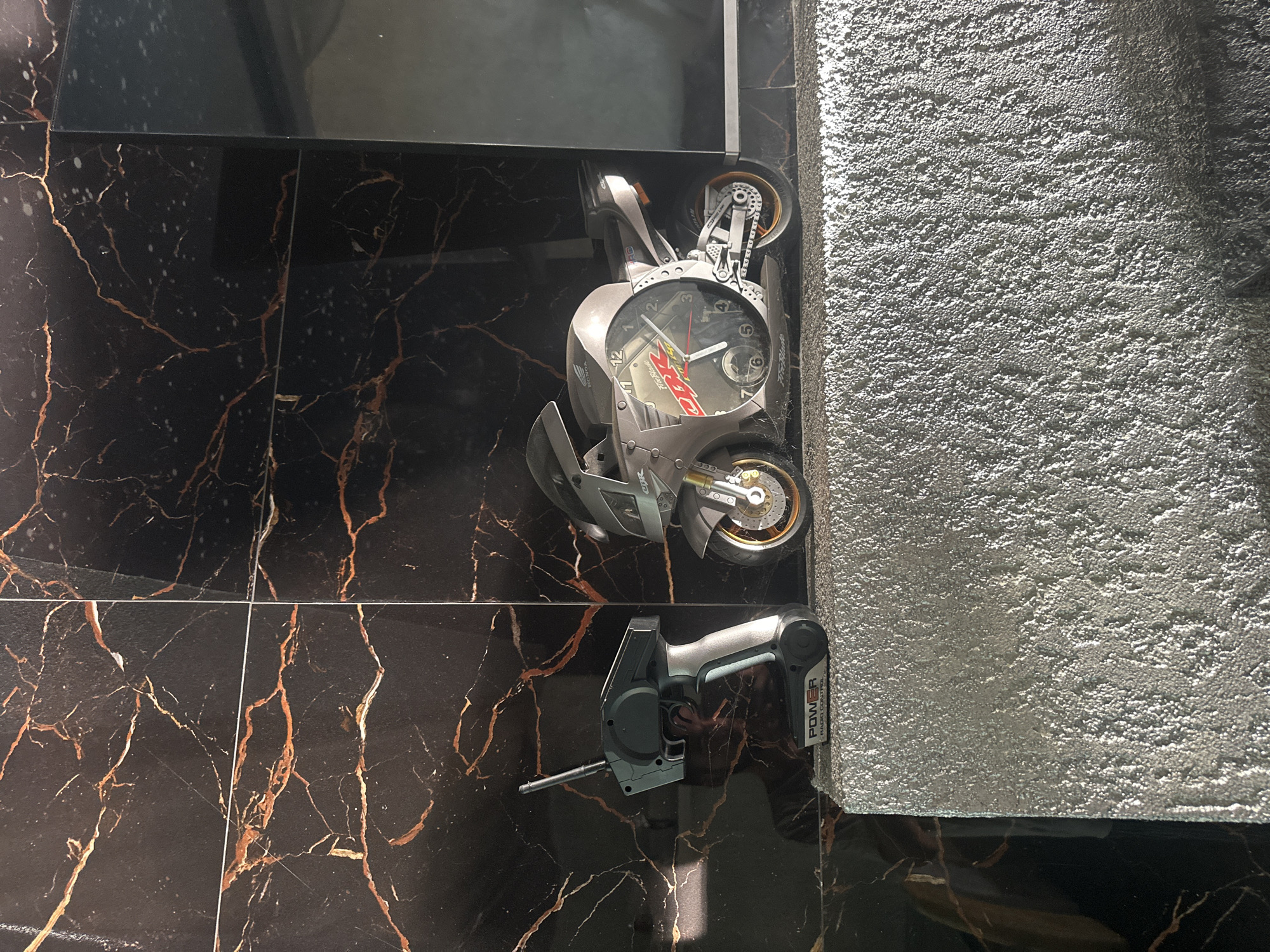 In Brca, Montenegro, on a textured silver shelf, a radio-controlled motorcycle clock sits next to its remote control. The motorcycle, a grey replica with gold accents, features a clock face embedded in its side. The remote control, also grey and silver, has an antenna. Behind the shelf, a black marble-patterned wall with thin gold veining serves as the backdrop. The lighting casts shadows and highlights the reflective surfaces.