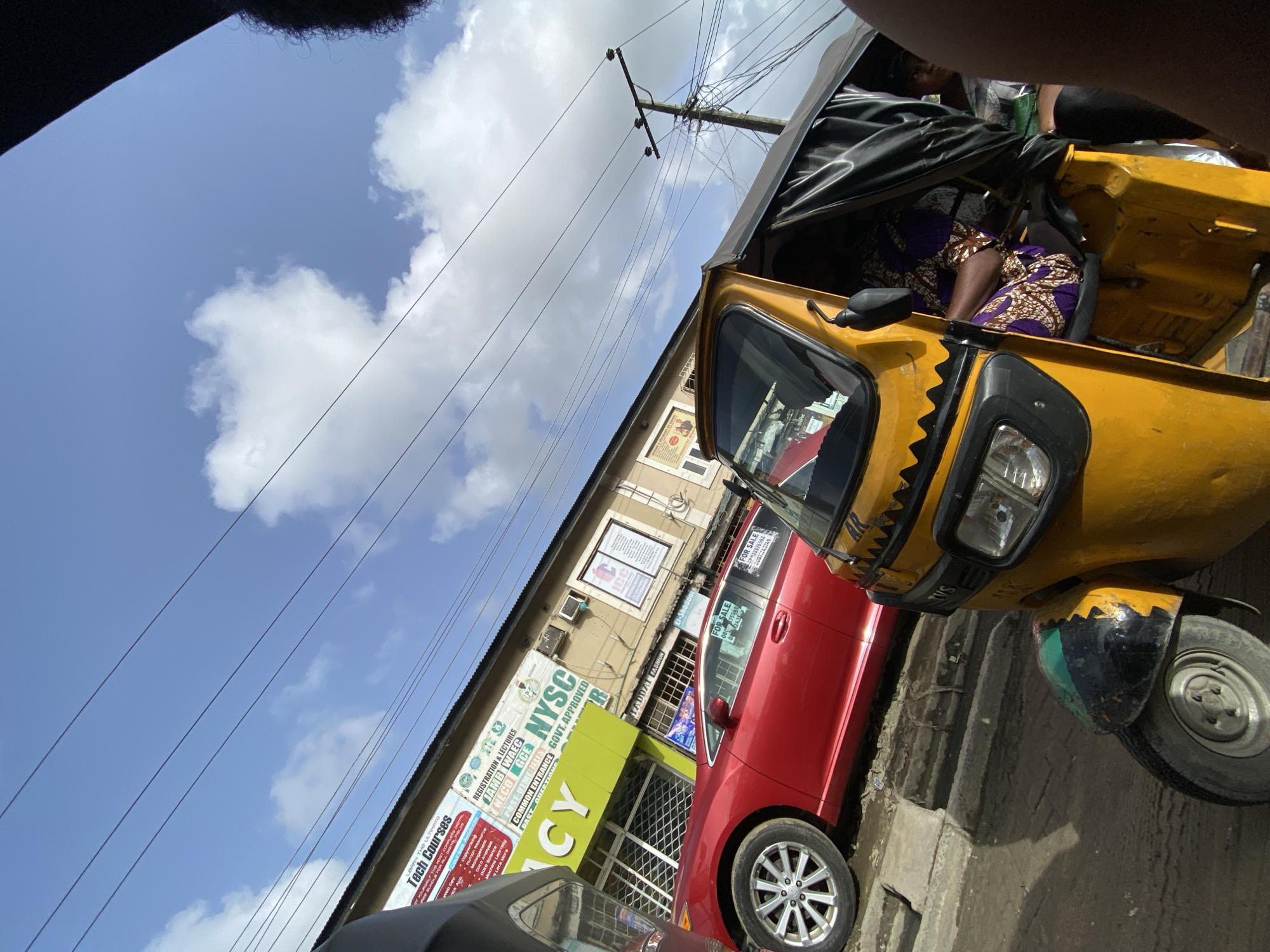 The image captures a street scene in Ado, Nigeria. A yellow "Keke Napep" (auto-rickshaw) is prominent on the right. Behind it is a red car, and adjacent to that is a building with signage indicating "Tech Courses," registration/lectures for JAMB, WAEC, GCE, NYSC, and common entrance examinations. The building also houses "El-Shadda TADDAL FASHION." Signs on the red car indicate it is for sale. An overhead wire runs across the top of the image against a backdrop of a blue sky with clouds. There appear to be people in the auto-rickshaw.