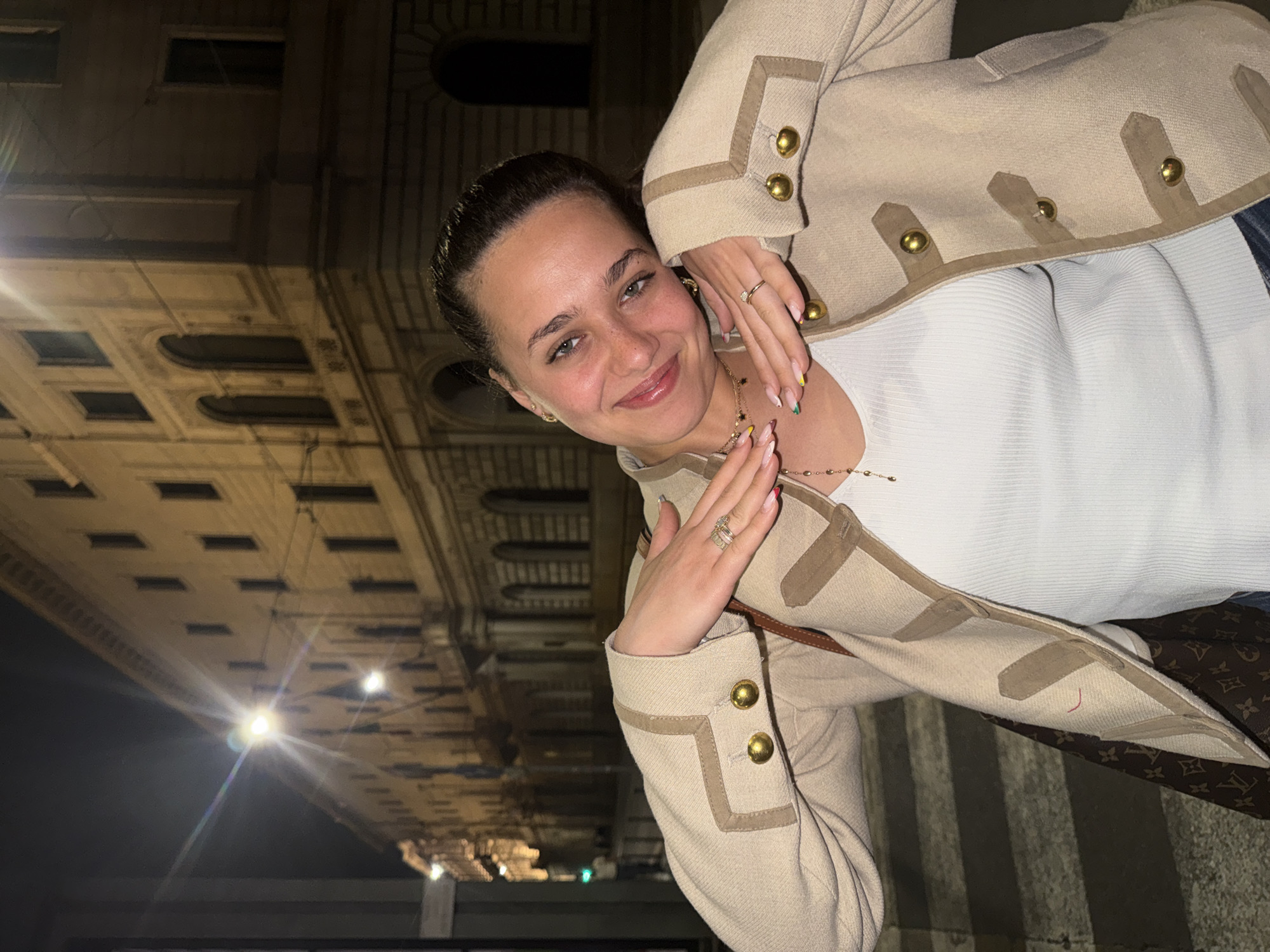 The media file captures a young woman posing for a photo outdoors at night in Milan, Italy. The image is taken at a tilted angle, with the woman's head resting towards the left side of the frame, looking directly at the viewer with a soft smile. She appears to be in her late teens or early twenties, with dark hair pulled back, light-colored eyes, and faint freckles across her face.
She is wearing a light beige or cream-colored jacket with tan trim and prominent gold-toned buttons on the front and cuffs. Beneath the jacket, a white ribbed top is visible. Her hands, adorned with rings (including one with a clear stone on her left ring finger), are positioned near her face and chest. Her fingernails are long and manicured, featuring colorful tips in shades of yellow, green, pink, and blue. A delicate gold-colored necklace with small charms hangs around her neck. On her right side (viewer's left), a portion of a bag with the distinctive brown Louis Vuitton monogram pattern is visible.
The background is dark, indicating nighttime, with artificial lights illuminating a classical-style building featuring ornate architectural details, such as arched windows and patterned cornices. One bright light source in the upper right portion of the background creates a starburst effect. The overall impression is a casual, happy moment captured in an urban setting at night.