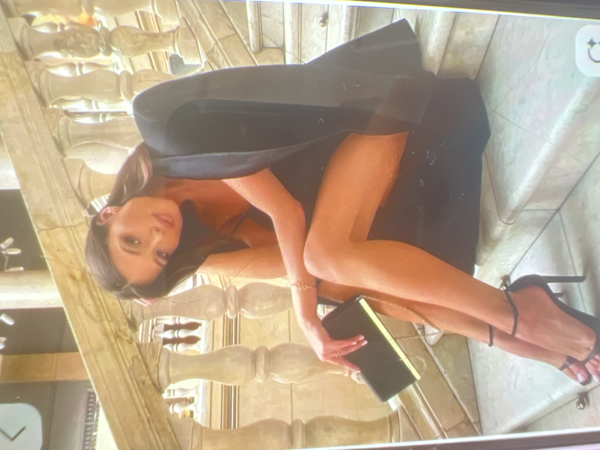 A woman in a black dress and blazer sits on marble stairs, holding a black clutch. Her legs are crossed, and she is wearing black heeled sandals. The stairs have ornate balustrades. The image quality is low and pixelated, suggesting it may be a screenshot of a video call or a low-resolution photograph. No location in Kara-Dıykan, Kyrgyzstan can be determined from the image.
