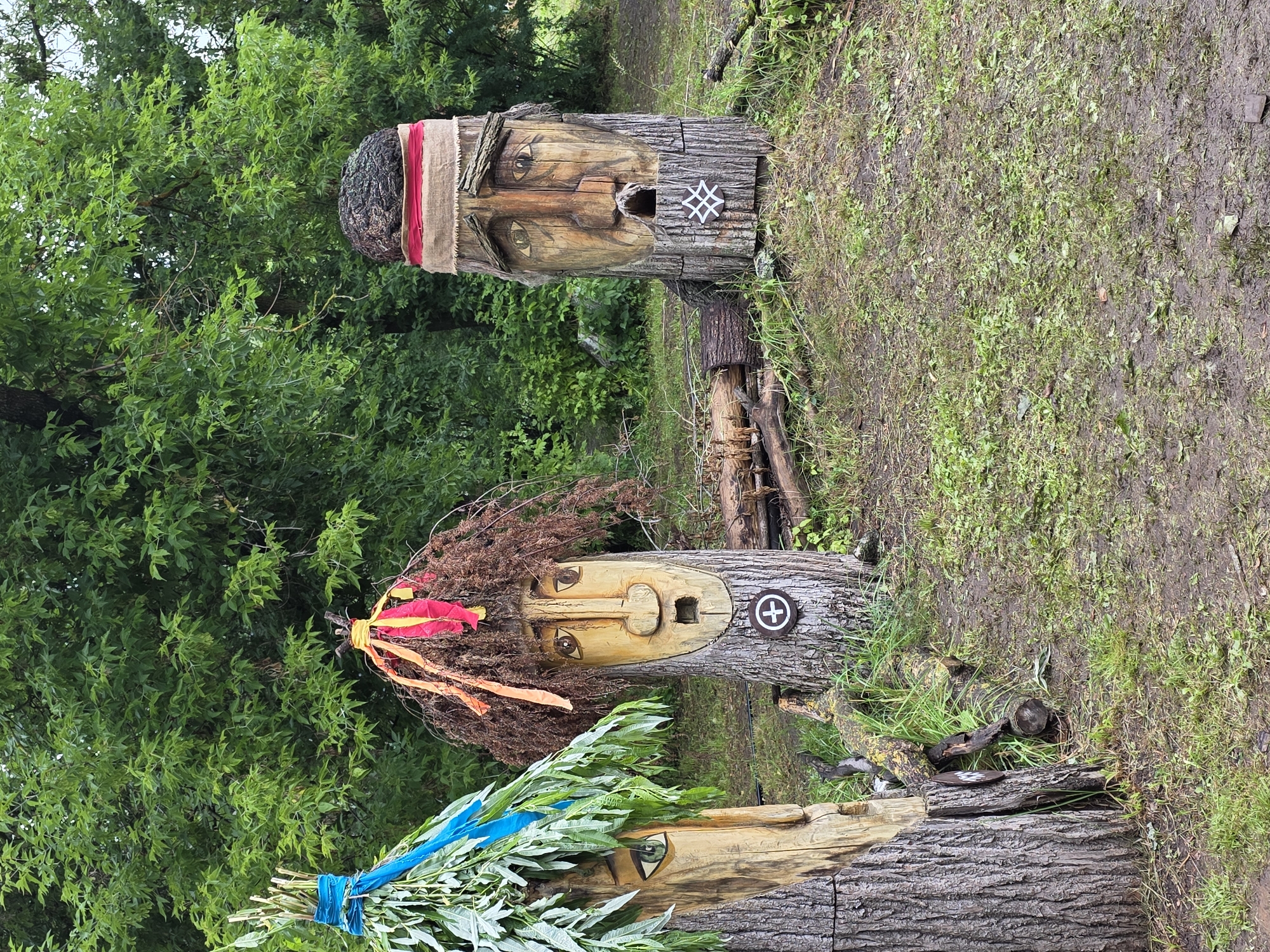 The image shows three carved wooden figures resembling faces, positioned outdoors in a grassy area with trees in the background. The figures are carved into tree trunks. The leftmost figure has a green eye and is adorned with green leaves and a blue ribbon. The central figure has a brown, bushy "hair" made of dried branches and is decorated with red and yellow ribbons. It has a carved face with an open mouth and a circular emblem with a cross inside, attached to its trunk. The rightmost figure has a rough, dark material on its head, resembling a cap, and is wrapped with a reddish-brown band and a lighter-colored fabric. This figure also has a carved face with an open mouth and a white symbol on its trunk. Fallen logs and branches are scattered around the base of the figures, and the ground is covered in grass and mud. The overall setting appears to be a park or a rural outdoor space. The location is identified as the village of Старые Быги (Starye Bygi), Russia.