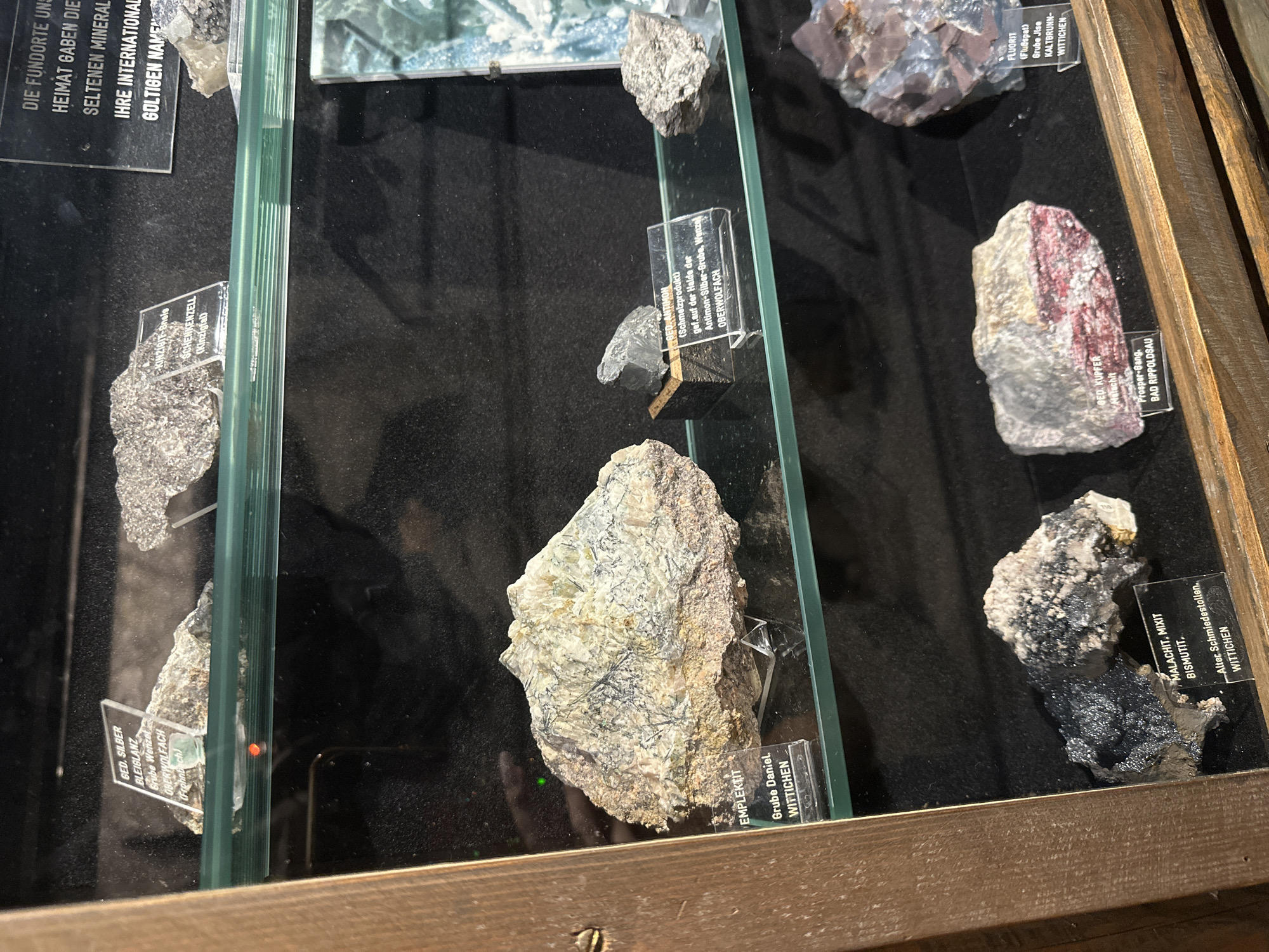 This image shows a display case with several geological specimens. The specimens are labeled with their names and origins. In the foreground, there is a dark, metallic-looking mineral specimen with a label that reads "MALACHIT, MIXIT, BISMUTIT, Alter Schmiedestollen, WITTICHEN". To the right of this, there is a pinkish-red mineral specimen labeled "GED. KUPFER, Malachit, Prosper-Gang, BAD RIPPOLDSAU". Above these, a large, light-colored rock specimen with dark veins is labeled "EMPLEKTIT, Grube Daniel, WITTICHEN". Further up, there are more specimens, including one labeled "GED. SILBER, BLEIGLANZ, Grube Wenzel, OBERWOLFACH". A black sign in the upper right corner reads "DIE FUNDORTE UND HEIMAT GABEN DIE SELTENEN MINERAL. IHRE INTERNATIONAL GOLTIGEN NAMEN". The display case is made of wood and glass. The lighting is dim, with some reflections on the glass. The image appears to be taken in a museum or exhibition. Schonach im Schwarzwald, Germany, is mentioned in the context of the origin of some of the minerals, specifically "WITTICHEN", which is a locality associated with mining in that region.