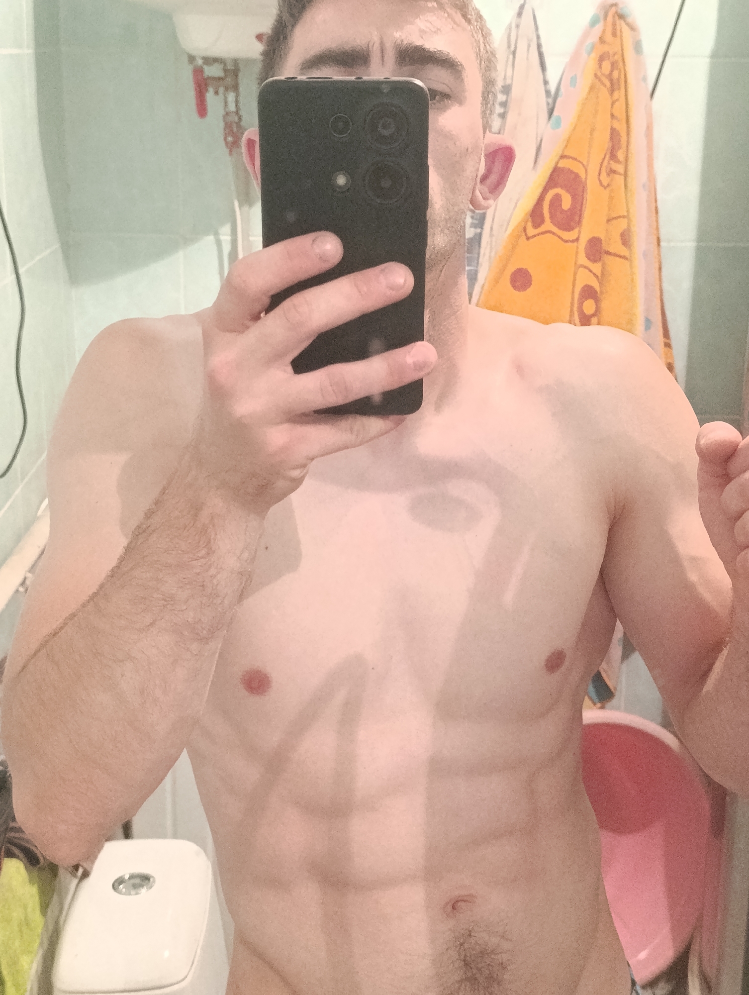 A man takes a mirror selfie in a bathroom. He is shirtless, showing his abs, and is holding a black smartphone in his right hand. The bathroom has light-colored tiled walls and a white toilet. A pink basin is visible in the background, as are two towels hanging on the wall. The image does not provide any specific details about the location in Fastiv, Ukraine.