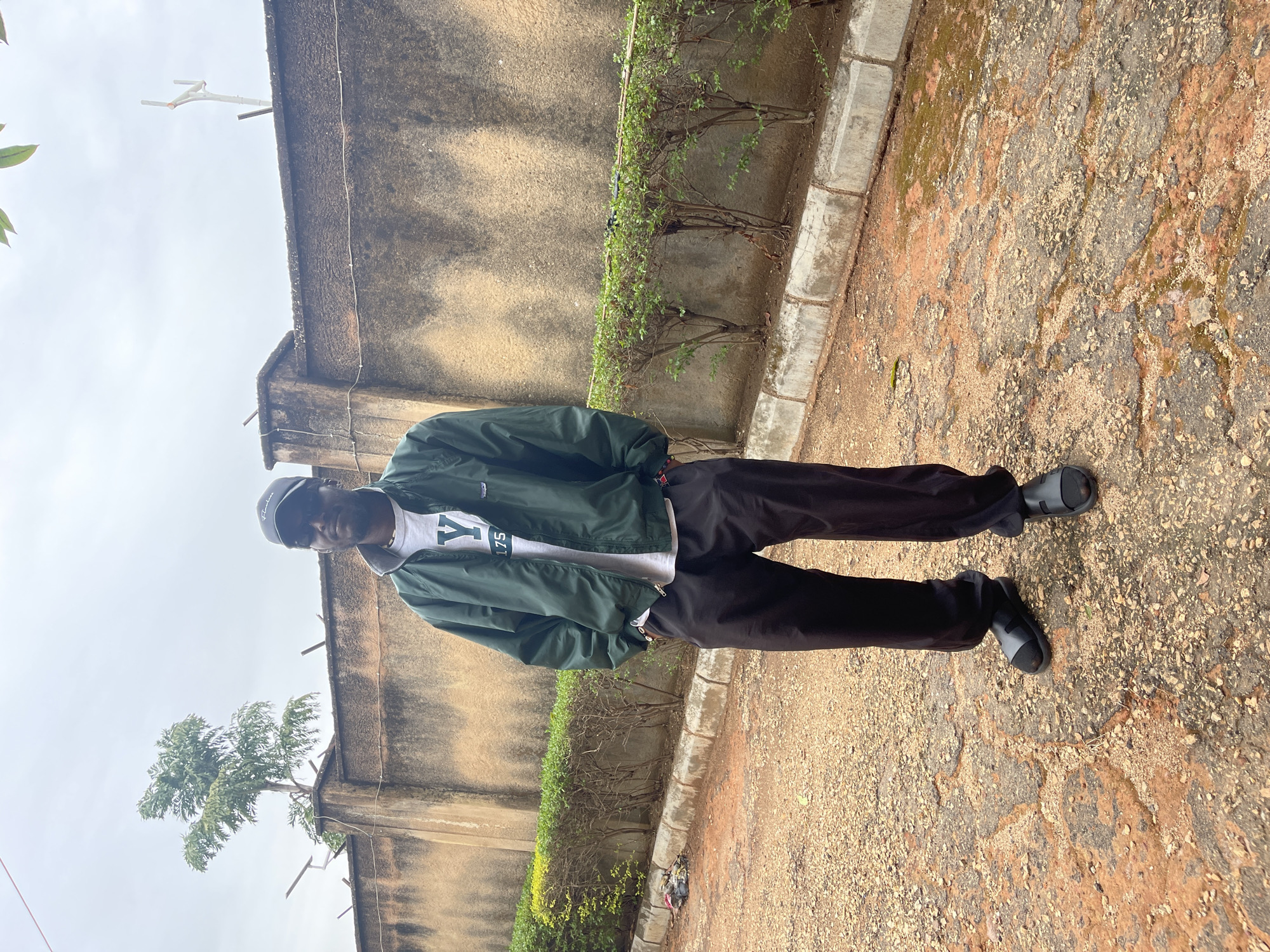A person is standing in an outdoor area. They are wearing a green jacket, a white t-shirt with a graphic, dark pants, and sandals. They also wear a baseball cap. The background features a weathered wall with a row of small shrubs at its base. The ground is a mix of dirt, small stones, and patches of what appears to be moss or discoloration. Given the presence of weathered walls and foliage, the location is likely in a residential area or a public space like a park or garden in the city of Jos, Nigeria.