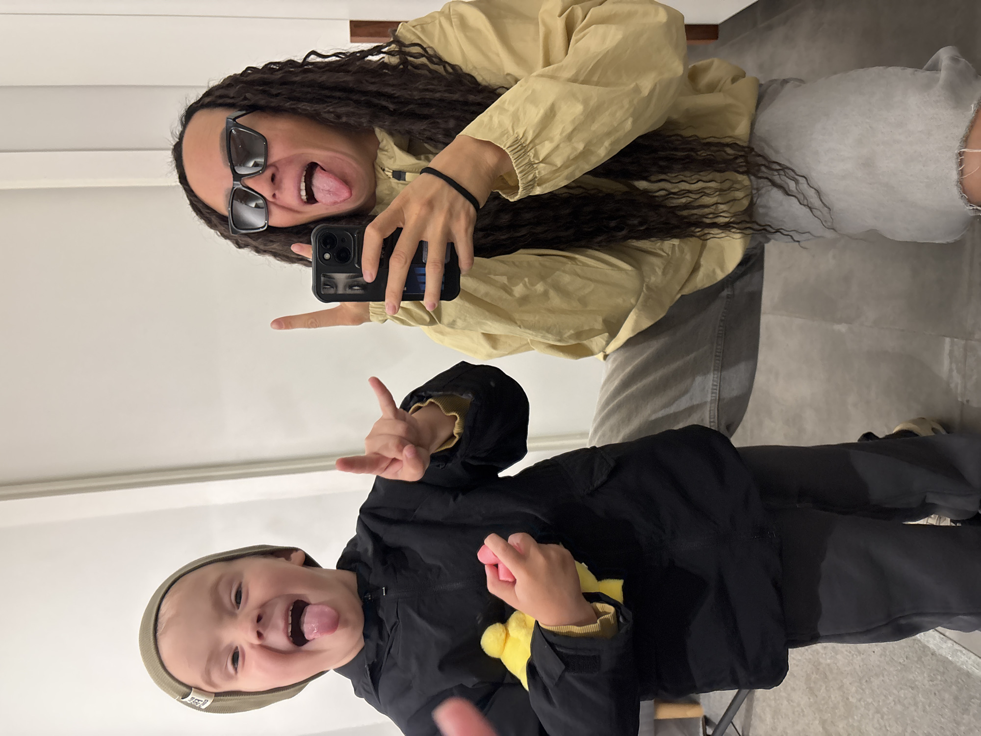 This is a playful mirror selfie featuring an adult and a young child, taken indoors in Chaikovsky, Russia. The image is oriented sideways, with both subjects making humorous expressions and gestures.
The adult, positioned in the upper part of the frame, has long, dark, wavy hair. They are wearing dark rectangular sunglasses, a light beige or tan windbreaker-style jacket, and light grey sweatpants. With their left hand, they hold a black smartphone with a dual-camera setup and a design depicting eyes on its case, appearing to be taking the selfie. Their right hand forms a "rock on" sign or a peace gesture. They are sticking their tongue out in a silly pose, indicating a cheerful mood. A black hair tie is visible on their right wrist.
The child, standing below the adult, is also making a funny face with their tongue out and a broad smile. They are wearing a beige or olive green beanie with a small white tag on it, a black hooded jacket with yellow lining visible at the cuffs, and dark grey pants. Their right hand is raised, mimicking the adult's "rock on" gesture. In their left hand, they hold a small, soft pink object, and a fluffy yellow item (possibly a toy or part of their clothing) is also visible near their arm.
The setting appears to be a brightly lit indoor space with white walls, featuring a horizontal white molding strip. The floor is a light grey, possibly concrete or tiled. The overall atmosphere conveyed by the subjects' expressions and gestures is one of joy, playfulness, and affection.