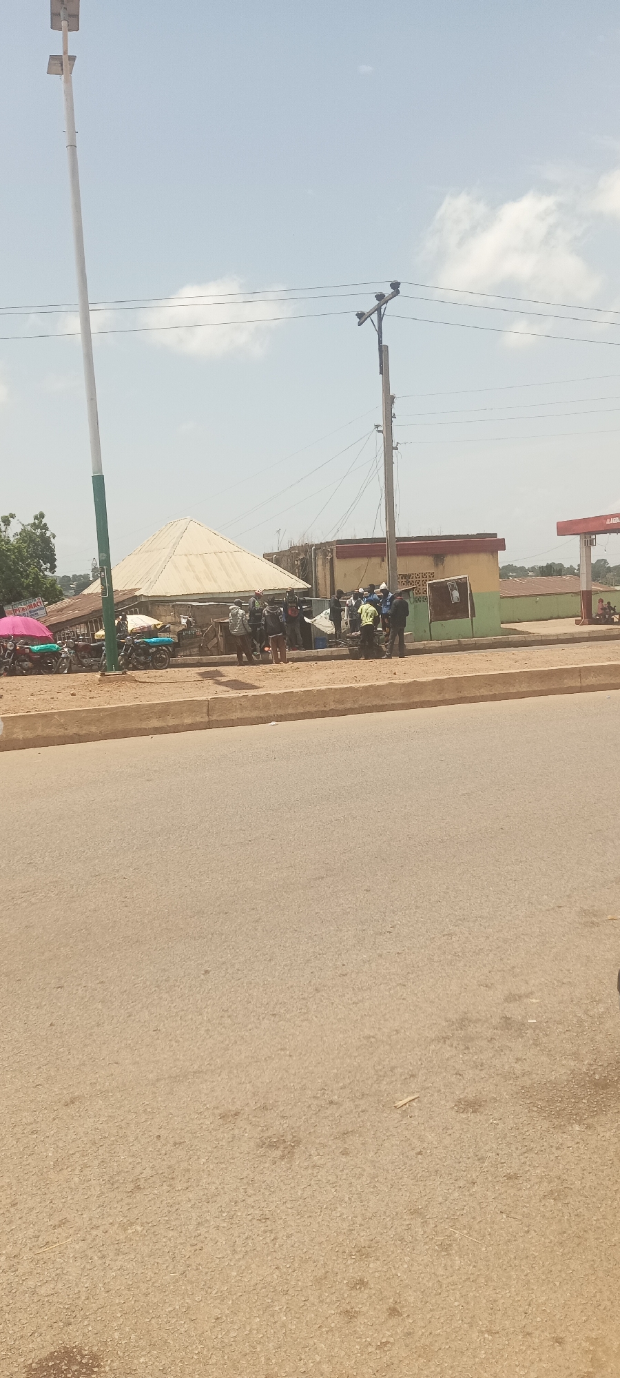 The image depicts an outdoor scene in Kuru Dungus, Nigeria, showing a street view during daylight. An asphalt road stretches into the foreground, flanked by a low concrete curb and a sandy shoulder.
A group of people stands gathered near a small, white-roofed structure resembling a market stall. Several motorcycles are parked nearby. Behind the group, there is a faded, tan-colored building with a green trim. Further in the background, a gas station with red and white signage is visible.
A tall, white streetlight pole with green accents stands on the left. Utility poles with power lines extend across the sky. The sky is partly cloudy.
