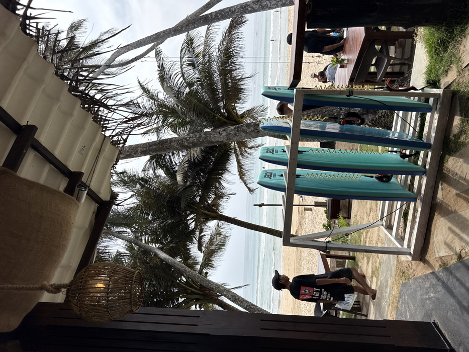The scene depicts a beachfront area in Dikwella, Sri Lanka, viewed from under a covered establishment. In the immediate foreground, a woven rattan light fixture hangs from a ceiling constructed with burlap-like material and wooden beams. A white security camera is mounted overhead. To the left, a dark wooden vertical pillar or door frame is visible.
In the midground, a male figure wearing a black cap, a black t-shirt with a white graphic, and dark shorts walks from left to right, carrying a white object. He appears to be barefoot or wearing flip-flops. Adjacent to him and slightly right, a white wooden rack holds four turquoise bodyboards, each branded with "JOCK SUPPLIES" and equipped with a yellow coiled leash. Further to the right, a dark wooden table with chairs is positioned. Several indistinct figures are present around this table; one male with lighter hair is partially visible behind the bodyboards, and another male in a dark shirt stands to the right.
The background reveals a sandy beach with numerous tall palm trees. The ocean stretches beyond the beach, exhibiting gentle waves. Several small human figures are visible in the distant water. The sky is overcast. The ground transitions from a compacted area to sand, with patches of grass near the establishment.