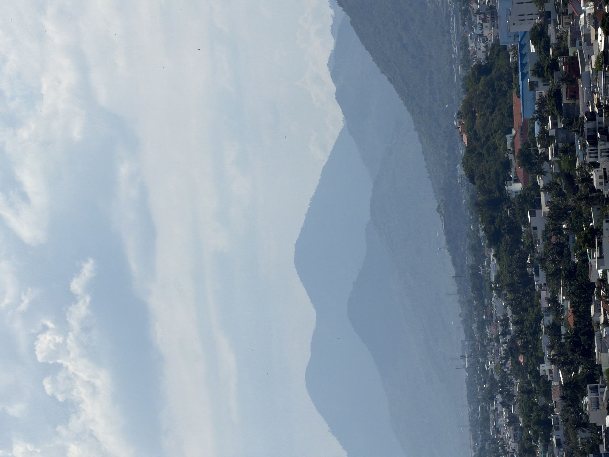 An aerial view captures a hazy, light-blue sky with scattered, wispy clouds. Below the sky, a series of layered, blue-toned mountains recede into the distance. The foreground features a densely populated urban landscape of Nha Trang Ward, Vietnam, with a mix of white and red-roofed buildings, interspersed with lush green trees, including palm trees. Numerous telecommunication towers are visible throughout the city. In the mid-ground, a prominent, tree-covered hill rises above the urban sprawl, topped with a structure. The overall scene suggests a sunny day with atmospheric haze.