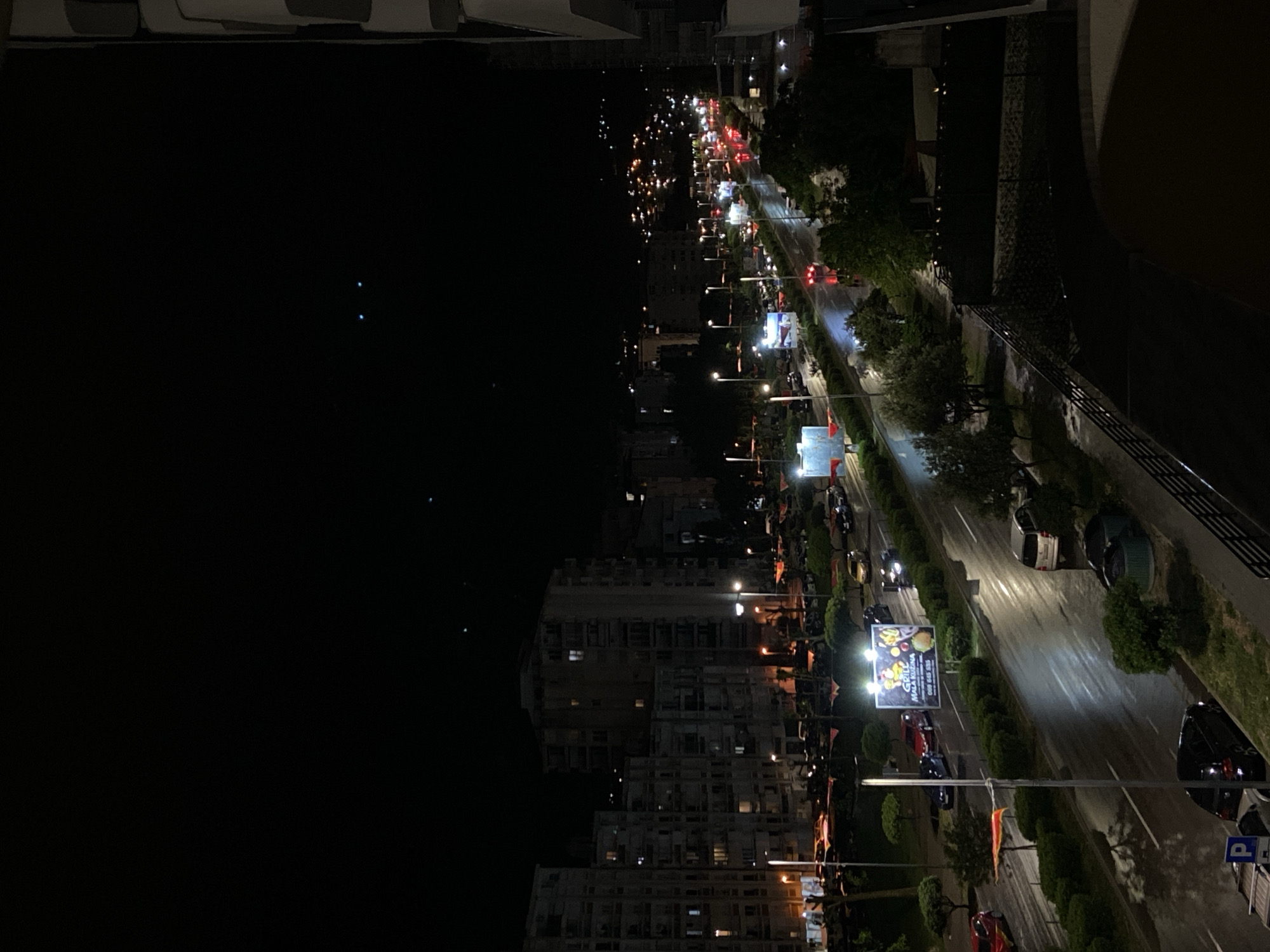 This is an elevated nighttime view of a street in Bar, Montenegro. The street is lined with trees and lamp posts. Buildings are visible to the left of the street. Cars are driving on the road. There is a billboard displaying food. The sky is dark.
