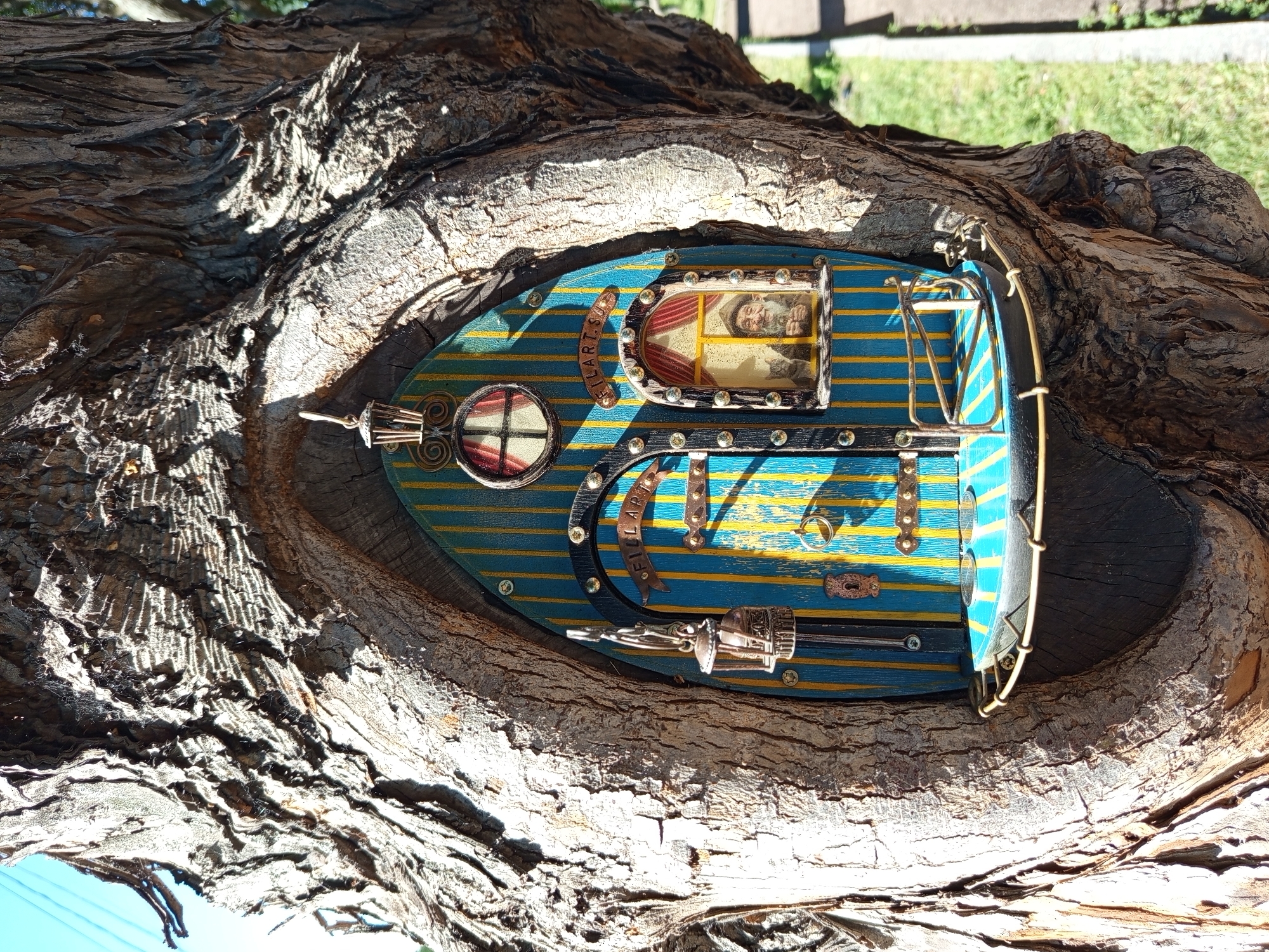 This media file captures a close-up, overhead view of a miniature, handcrafted boat nestled within a hollowed-out section of a tree trunk. The boat is brightly colored with blue and yellow stripes, adorned with metallic embellishments and small screws. A circular window with red-lined panes is visible on the left side of the boat, and a rectangular framed image of what appears to be a man in traditional attire is positioned in the center. The word "SELAR.SU" is inscribed in a curved metallic piece on the boat's surface.
The tree trunk's rough, textured bark dominates the foreground and background, with strong sunlight casting dappled shadows across its surface, suggesting it is daytime and likely sunny. The boat is the clear focal point, seemingly placed as a decorative or artistic element within the natural setting of the tree. The broader scene beyond the tree suggests an outdoor environment, with hints of greenery and a paved area in the distance, indicative of a park or garden. The overall impression is one of whimsical artistry integrated into nature. The location context of Saint Petersburg, Russia, is provided but not visually evident within the image itself.