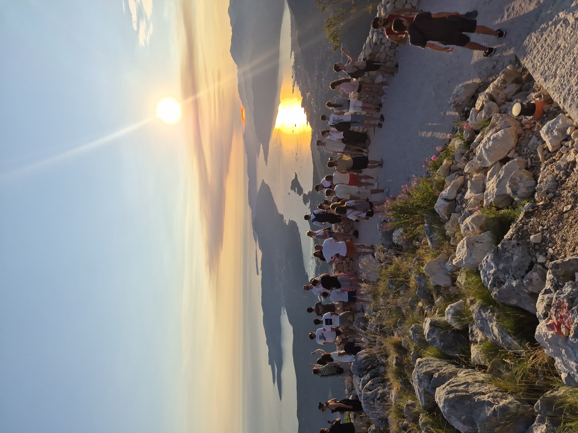 A crowd of approximately 30 people are gathered on a rocky, elevated vantage point overlooking a body of water and distant mountains during sunset. The sun is low in the sky, casting a warm, golden glow across the water and illuminating the clouds. Several individuals are standing, while others are seated on a low stone wall. The people are diverse in age and gender, dressed in casual clothing. Some are taking photographs with their phones, while others are looking out at the view. The environment is rocky and arid, with sparse vegetation. In the distance, a bay or fjord is visible, with hills and mountains rising on either side. The city of Kuk, Montenegro, is situated along the coastline in the distance, characterized by its lights reflecting on the water. The overall impression is of a scenic viewpoint where tourists are enjoying the sunset.