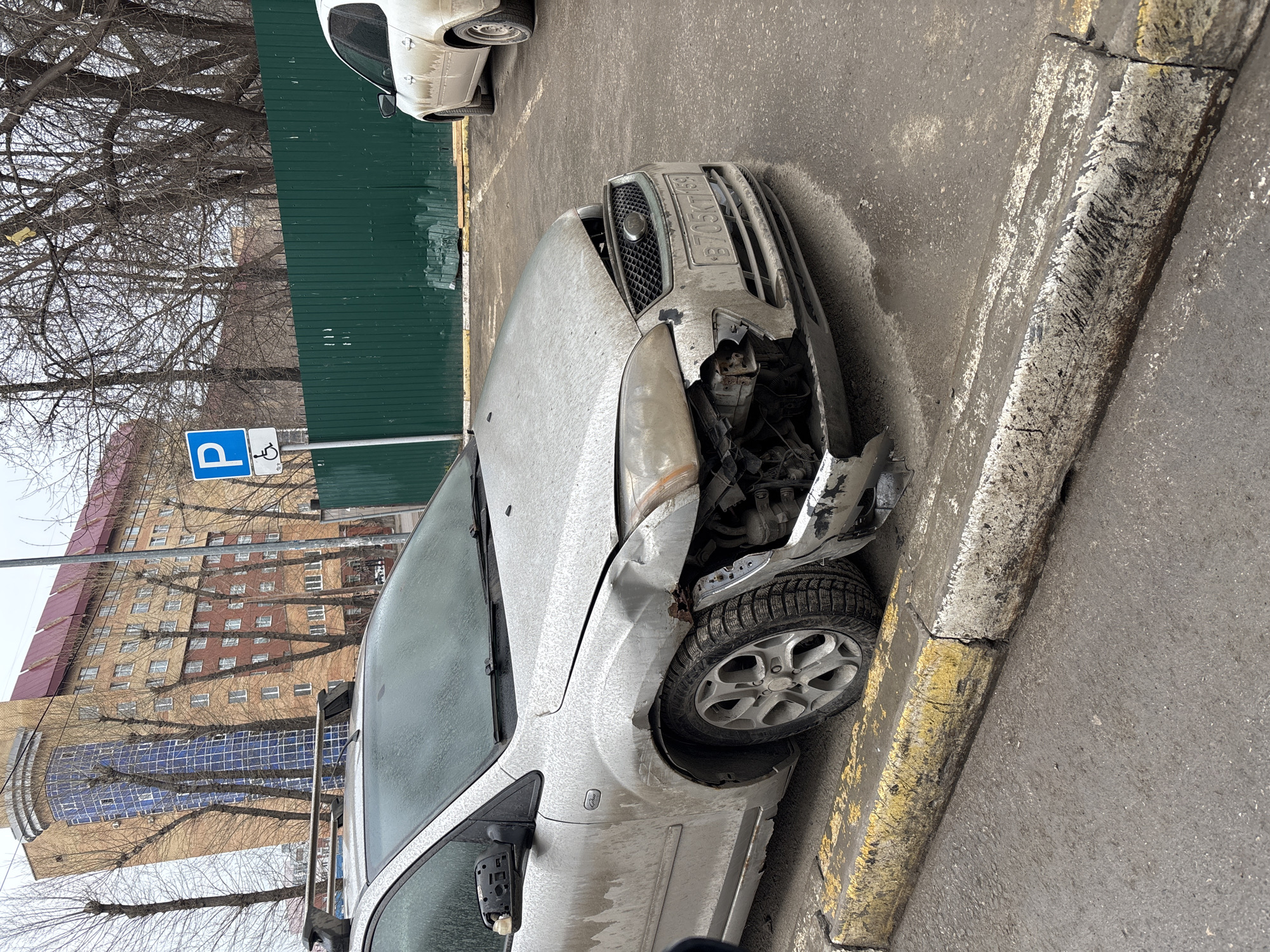 The image shows a damaged silver Ford car parked next to a curb in a city. The front of the car is heavily damaged, particularly on the passenger side, with missing parts and visible damage to the headlight, bumper, and front panel. The car's license plate, partially visible, reads "B705KT 159". The car is positioned on a street with a sidewalk and a parking space sign indicating disabled parking. In the background is a multi-story brick building with many windows. Bare trees are visible between the parking sign and the building. A green fence is also visible, partially obscuring the scene. The overall impression is that the vehicle has been involved in a collision. Based on the license plate (region code 159), the city is Perm, Russia.