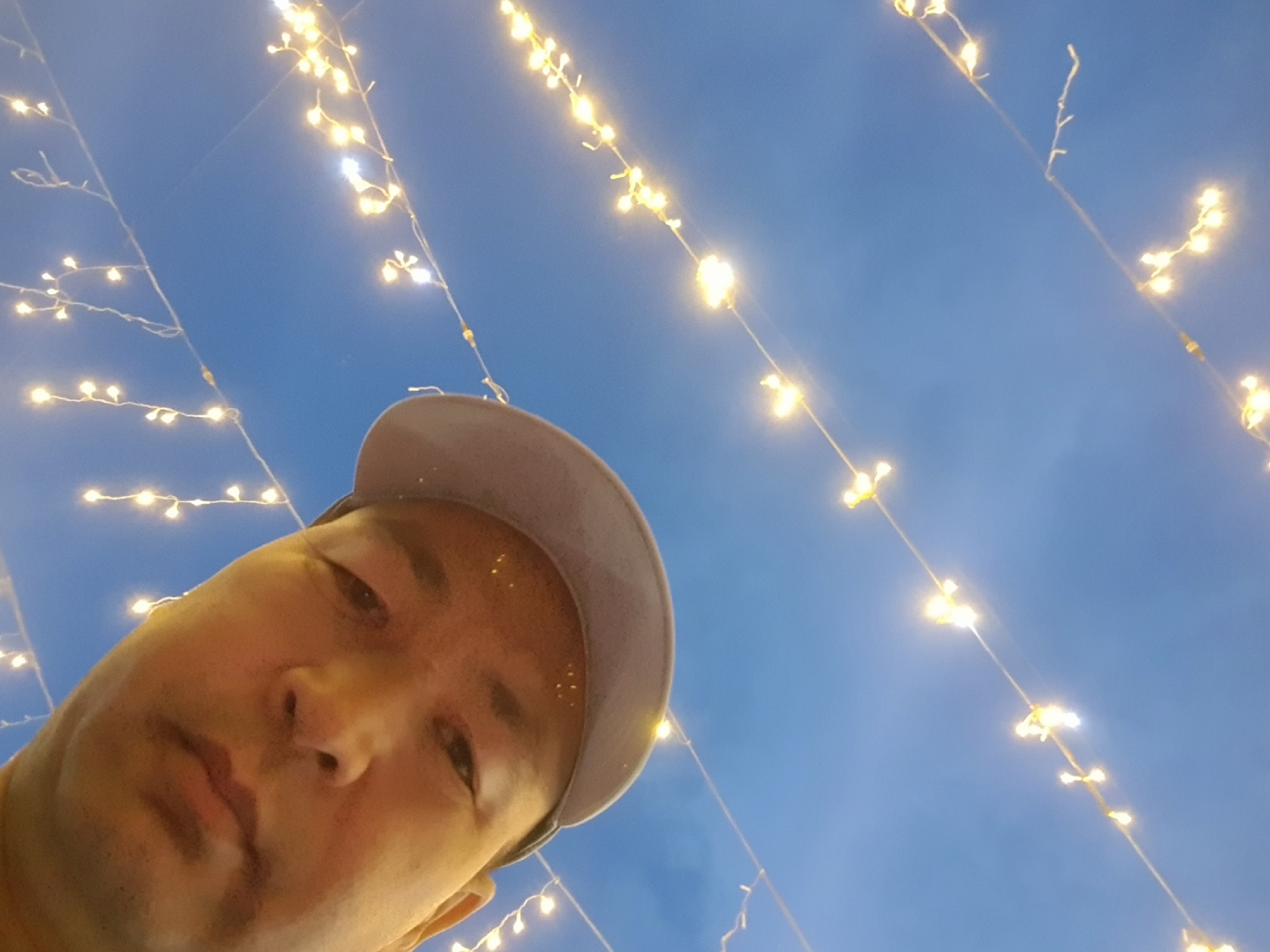 This media file is a low-angle selfie taken by a person wearing a light-colored baseball cap. The subject's face, partially obscured by the cap, is looking upwards towards a deep blue sky. The sky is adorned with strings of small, warm-toned fairy lights that are illuminated, creating a festive or decorative atmosphere. These lights are strung across the frame at various angles, with some appearing closer and brighter than others. The overall impression is that of an outdoor setting, likely at dusk or night, given the illuminated lights against the darkening sky. The location context is Kurchatov, Kazakhstan, which might suggest a possible event or holiday celebration where such decorations would be used. No text is visible in the image. The image is focused on the person's face and the overhead lights, suggesting a casual and personal moment captured by the individual.