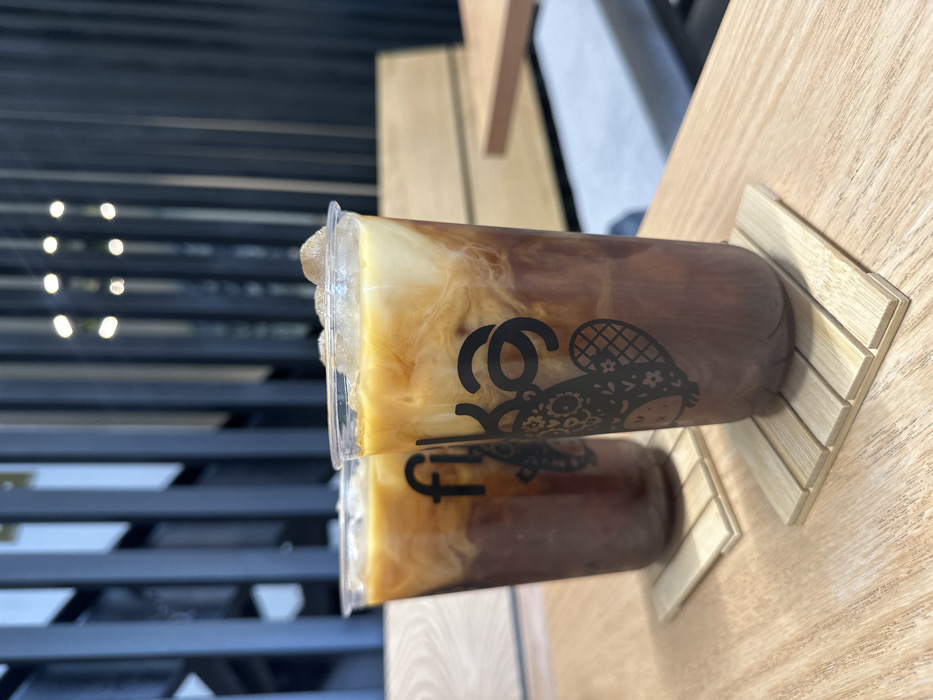 Two plastic cups filled with iced coffee sit on wooden coasters. Cream swirls within the coffee, creating a marbled effect. A black logo, featuring a stylized character, is printed on the cups. The cups are positioned side-by-side, with a portion of each cup visible. The background includes a blurred view of a window with horizontal slats and a light wooden surface.