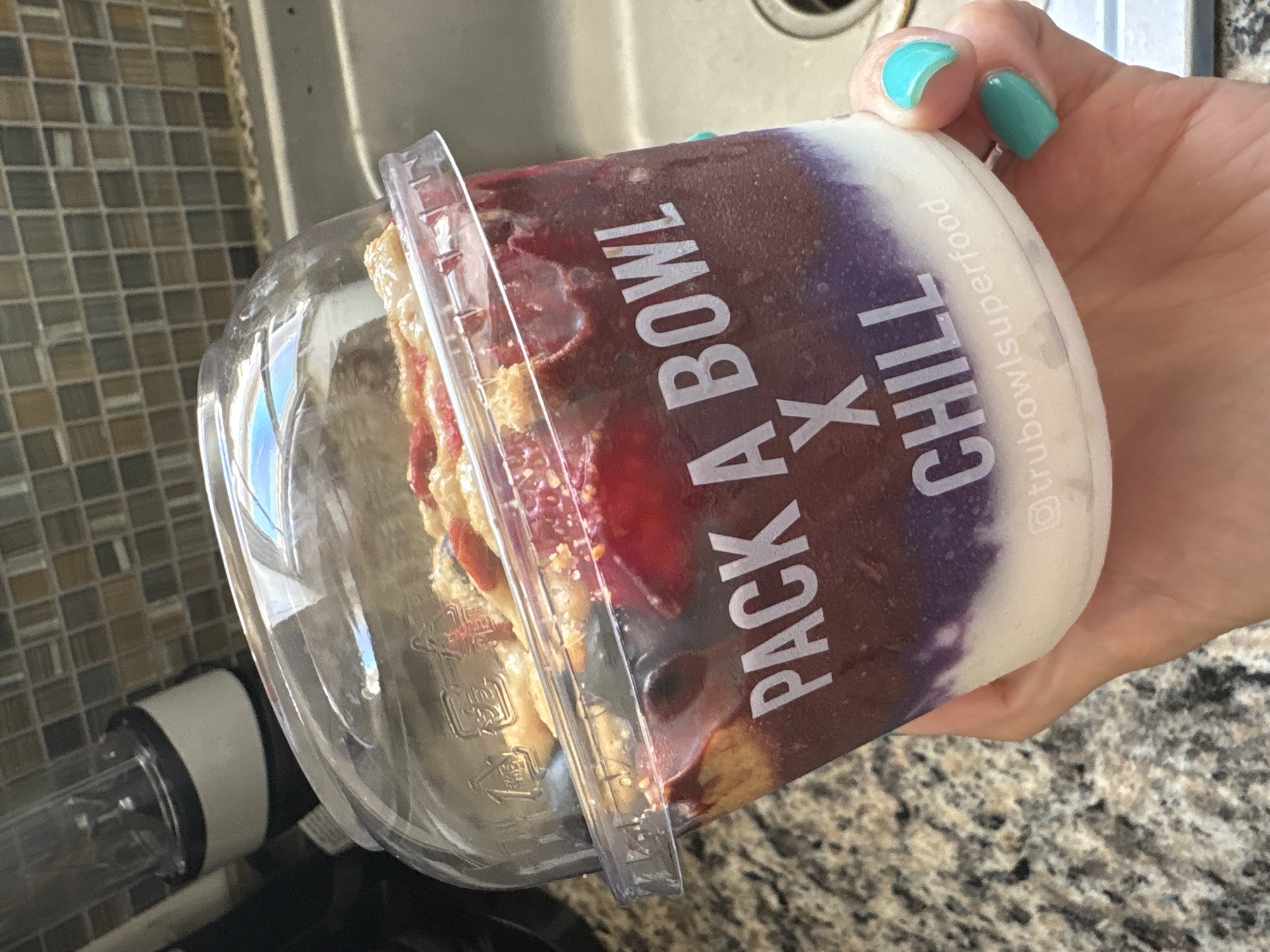 A hand with turquoise nail polish holds a clear plastic container of a "PACK A BOWL X CHILL" smoothie bowl by "trubowlsuperfood" over a brown speckled countertop in a kitchen, with the edge of a sink and a small appliance visible in the background. Based on the company name on the container, the location is Seal Beach, United States of America, where the company is located. The bowl appears to be multi-layered with various food items.