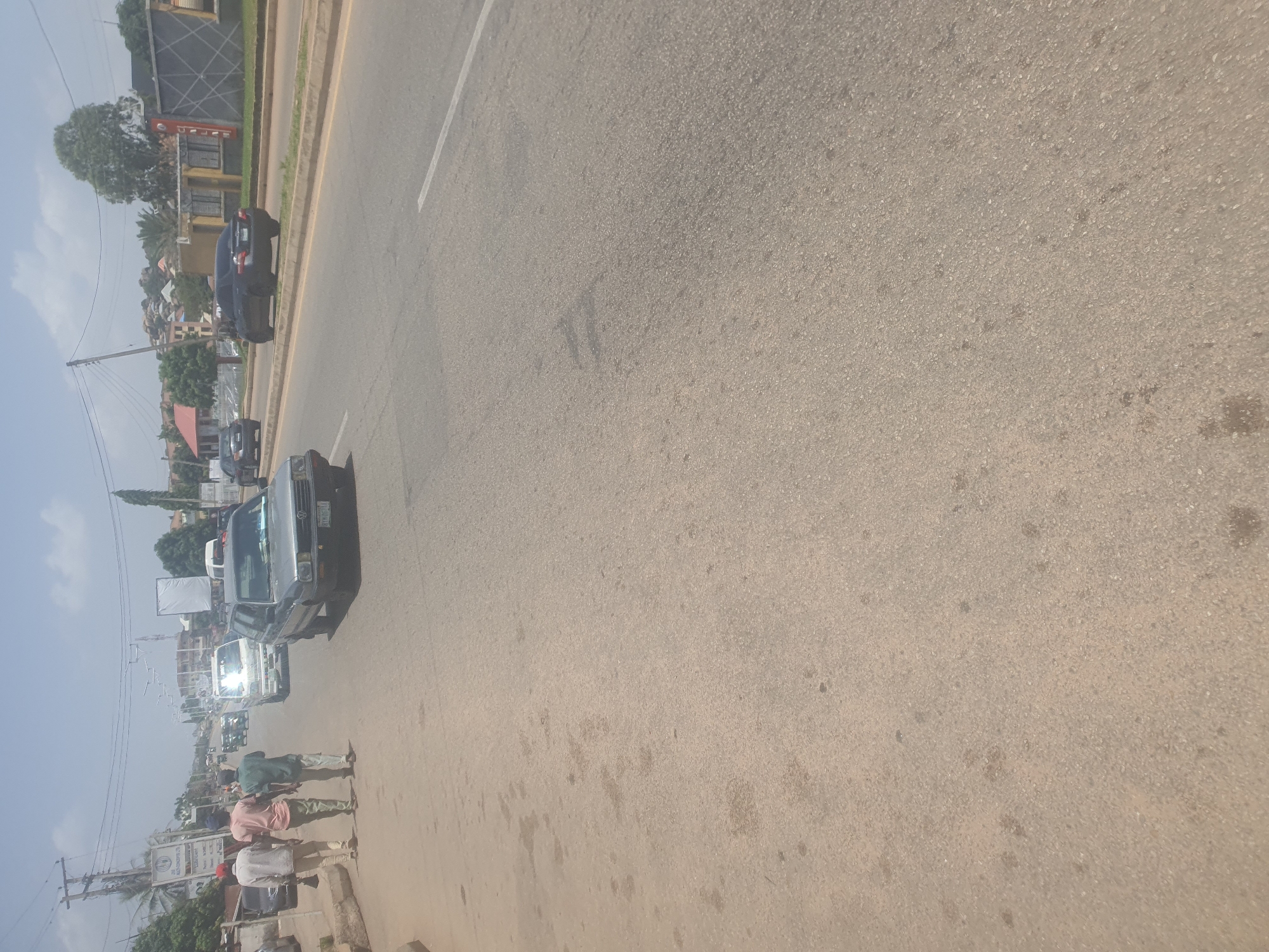 In New Stadium, Nigeria, a street scene unfolds under a bright, hazy sky. A grey Volkswagen Golf sedan is positioned on the right lane of a paved road. To the left of the road, three men are walking on a concrete sidewalk. The man in the front wears a white long-sleeved shirt and dark trousers, carrying a stick. The middle man is in a pink t-shirt and light green trousers, and the third man is in a green shirt and light-colored trousers. Further down the road, various other vehicles are visible, including a white mini-bus and a dark-colored sedan. Buildings line the right side of the road, with some featuring signs and architectural details. The environment is typical of a developing urban area, with overhead power lines and streetlights.