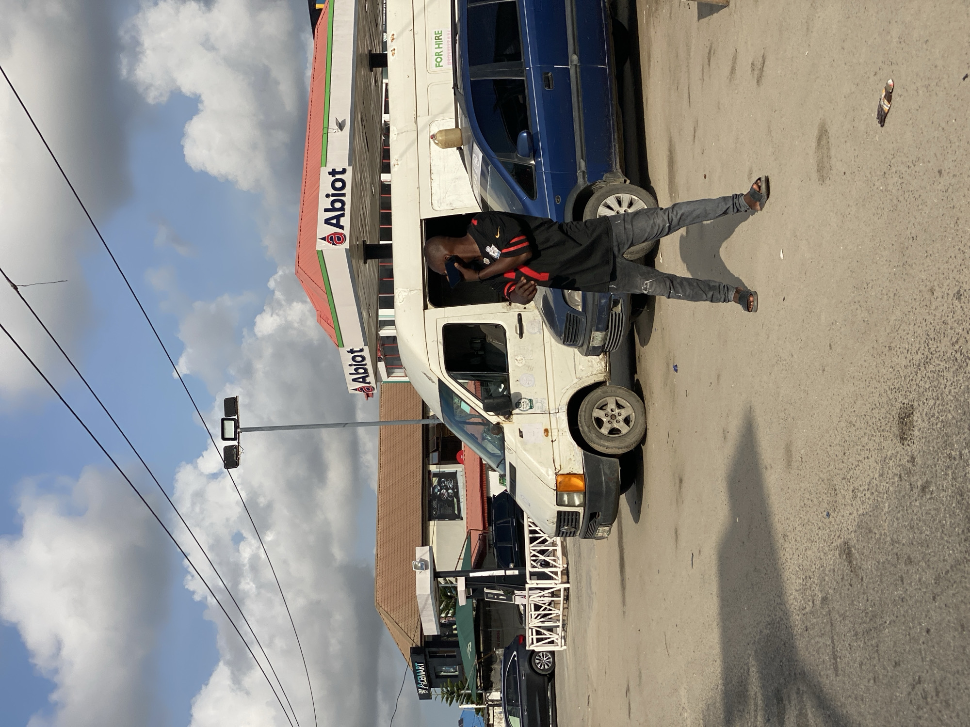 The image depicts a man standing in the open driver's side door of a white van, seemingly on a phone call, at an Abiot gas station in Ikorodu, Nigeria. A blue car is parked adjacent to the van, partially obstructing the view of the Abiot-branded gas station. Power lines stretch across the sky filled with cumulus clouds. The ground is paved and a sandal rests on the road. Other parked vehicles and shops are visible in the background.
