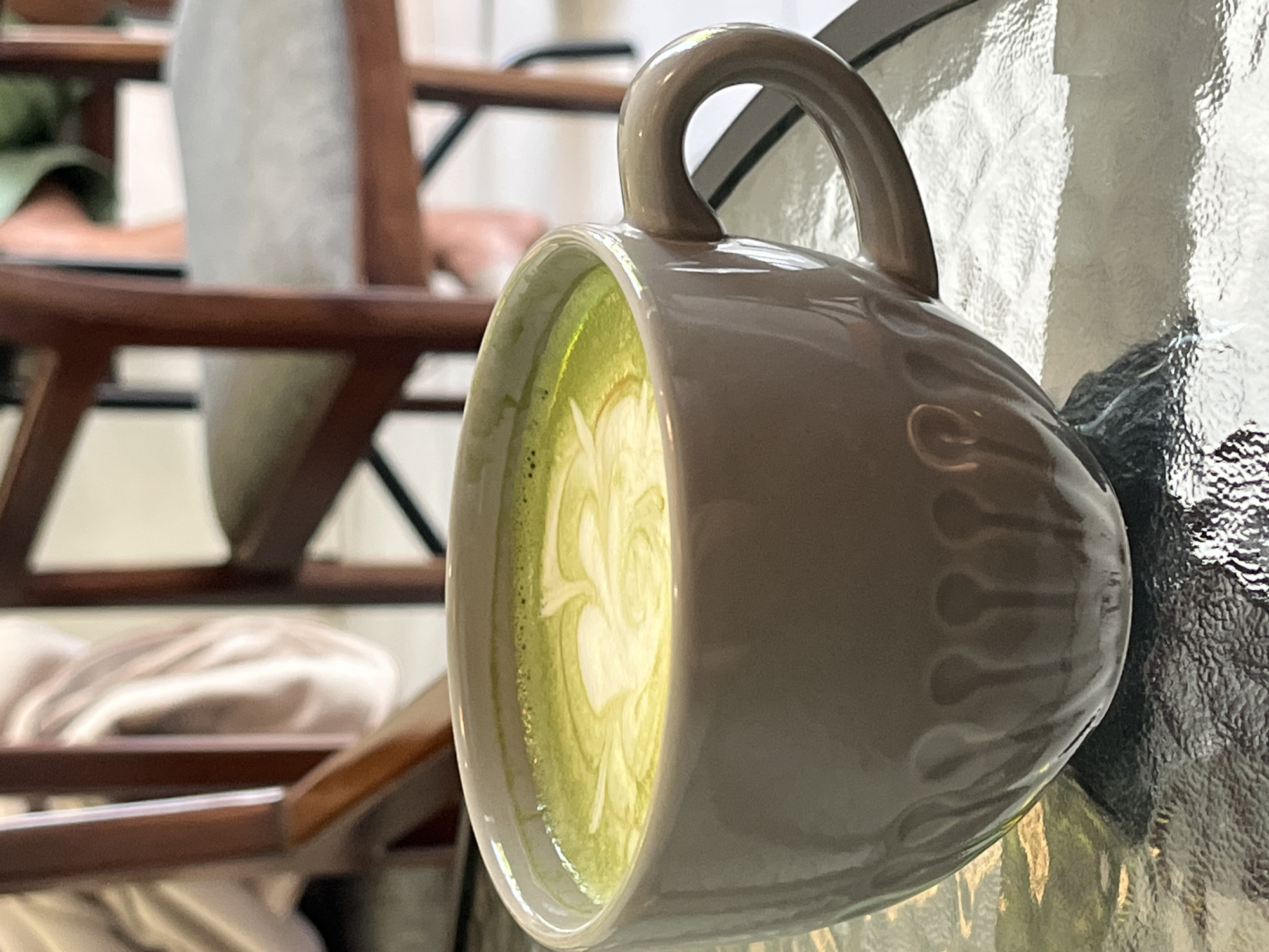 A close-up shot depicts a light-gray mug filled with a matcha latte, showcasing latte art on its surface. The mug rests on a textured glass surface, possibly a table. In the blurred background, wooden chairs with cushioned seats are visible. The location is unknown.