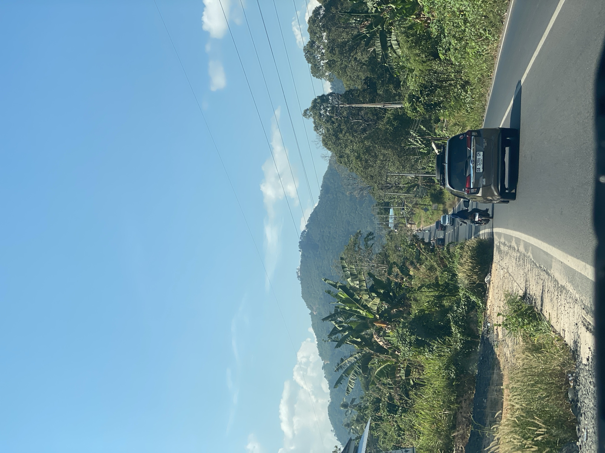 The image depicts a road in the city of Padang Panjang, Indonesia, surrounded by lush green vegetation and rolling hills. The sky is a clear blue with scattered white clouds. In the foreground, a paved road with a white line marking the lanes is visible, with a gray car driving away from the camera. To the left of the road, there is a patch of dry grass and some foliage, while to the right, dense green trees and banana plants line the roadside. In the distance, a mountain covered in trees can be seen. Several other vehicles, including a motorcycle with a rider, are also on the road. Power lines stretch across the sky, supported by utility poles. The scene appears to be a rural or semi-rural area within Padang Panjang.