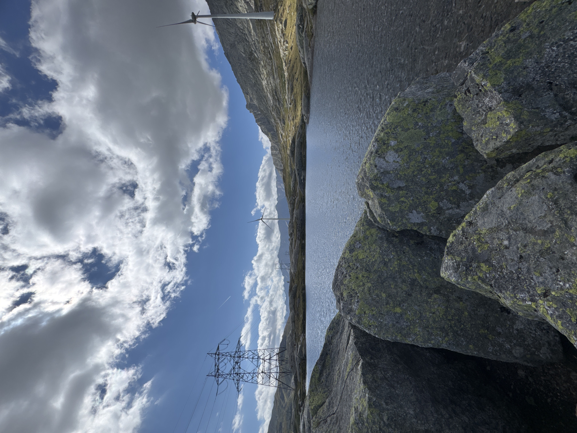 The image depicts a mountainous landscape near Airolo, Switzerland, characterized by a small lake in the foreground, surrounded by rocky terrain. In the distance, several wind turbines are visible, suggesting a renewable energy installation. A prominent high-voltage power line tower with accompanying electrical cables stands to the left of the frame. The sky is partly cloudy, with a mixture of blue sky and white, gray clouds. The rocks in the foreground are large and appear to be covered with lichen, adding texture to the scene. The lake's surface is rippled, indicating a slight breeze. The overall atmosphere is natural and rugged, with human infrastructure integrated into the landscape. There are no people visible in the image.