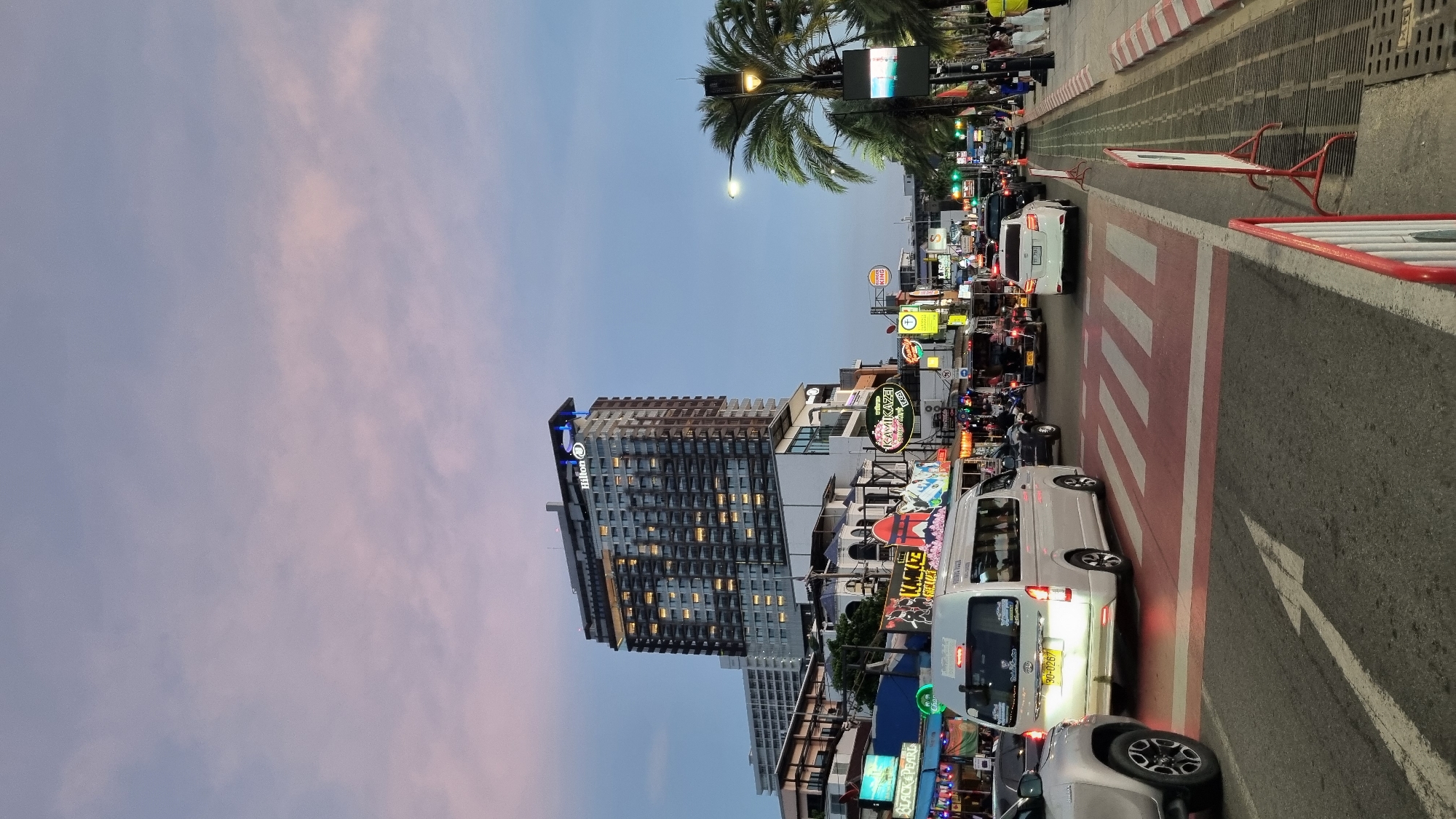 The image depicts a busy street scene in Pattaya City, Thailand, at dusk. A tall Hilton hotel dominates the skyline, its facade illuminated by numerous windows. Below the hotel, a vibrant street is lined with businesses, characterized by colorful neon signs, including "Kamikaze" and "Black & Pearl." The street is congested with traffic, featuring a variety of vehicles such as cars, vans, and motorbikes. Pedestrians can be seen on the sidewalks, and palm trees line the right side of the street. A pedestrian crossing with red and white markings is prominent in the foreground, with cars and a white van waiting at the intersection. The sky above displays a soft gradient of pink and blue, indicative of sunset. The overall atmosphere is one of urban activity and commercial vibrancy.
