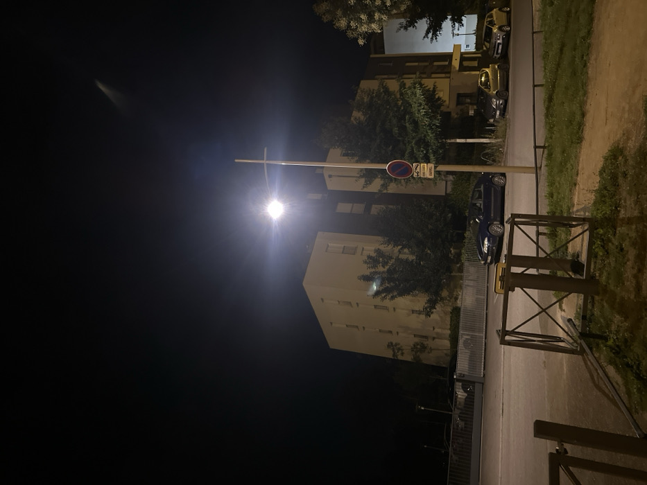 This is a nighttime, static view of a street scene in Strasbourg, France. The dark sky dominates the upper portion of the image, with a faint, diagonal light streak visible in the upper right. A prominent, bright streetlamp on a tall, light-colored pole illuminates the midground, causing significant lens flare. A red circular "no parking/stopping" sign with a smaller white rectangular sign below it is affixed to the streetlamp pole.
In the foreground, a section of an asphalt road is visible, bordered by a grassy patch on the right where a metal barrier or frame lies partially on the ground. The midground features the asphalt road, with multiple vehicles parked along its sides. A dark blue sedan is parked on the right side of the road, facing left. Further down the road on the right, two compact cars, one yellow/lime green and one beige, are parked facing right. On the left side of the road, behind a metal gate or fence, several dark vehicles are parked.
The background consists of multi-story residential or commercial buildings. To the left, a light-colored building with multiple windows and balconies rises. To the right and behind the streetlamp, darker buildings are partially visible. Trees with dense green foliage are dispersed throughout the background, between the buildings and along the roadside. No individuals are present in the scene, and no actions or interactions are depicted.