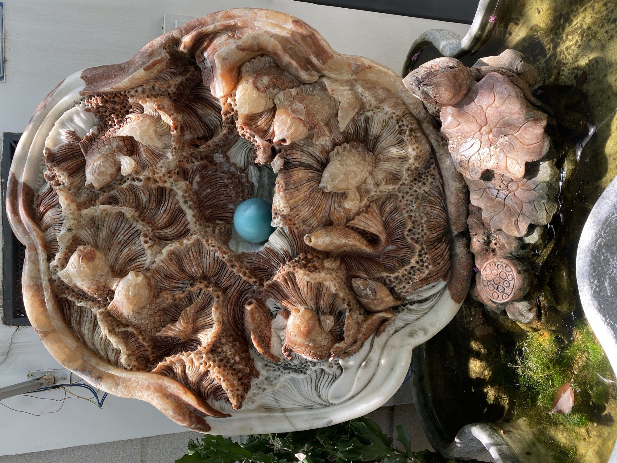 This image shows an ornate, round stone carving featuring multiple fish and waves. The carving is made of marble with reddish-brown and white veins. In the center of the carving, a bright blue sphere is embedded. The sculpture is placed in a stone basin filled with water, where aquatic plants and a fallen leaf are visible. The sculpture appears to be part of a garden or outdoor decorative feature. The exact location within Đà Nẵng, Vietnam is not discernible from the image.