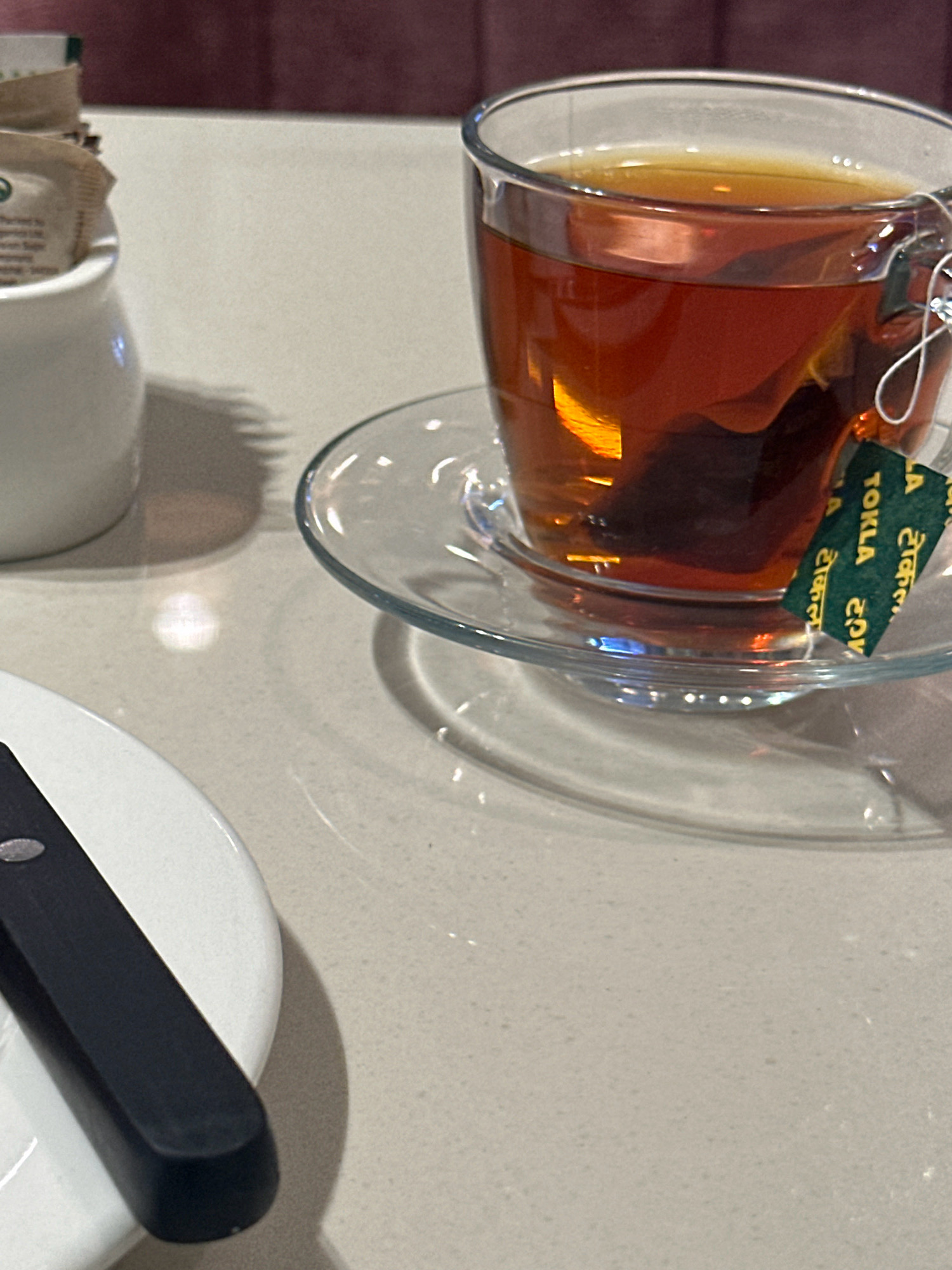 A close-up, eye-level shot shows a clear glass mug filled with amber-colored tea, with a tea bag submerged within. The mug rests on a clear glass saucer. A green tag attached to the tea bag displays the word "TOKLA" in English and what appears to be Nepali script. To the left, a white ceramic sugar dispenser contains packets of sugar. A black knife lies on a white plate in the foreground. The scene is set on a light-colored countertop. The background is blurred, showing a dark red surface. This image was taken in Kathmandu Metropolitan City, Nepal.