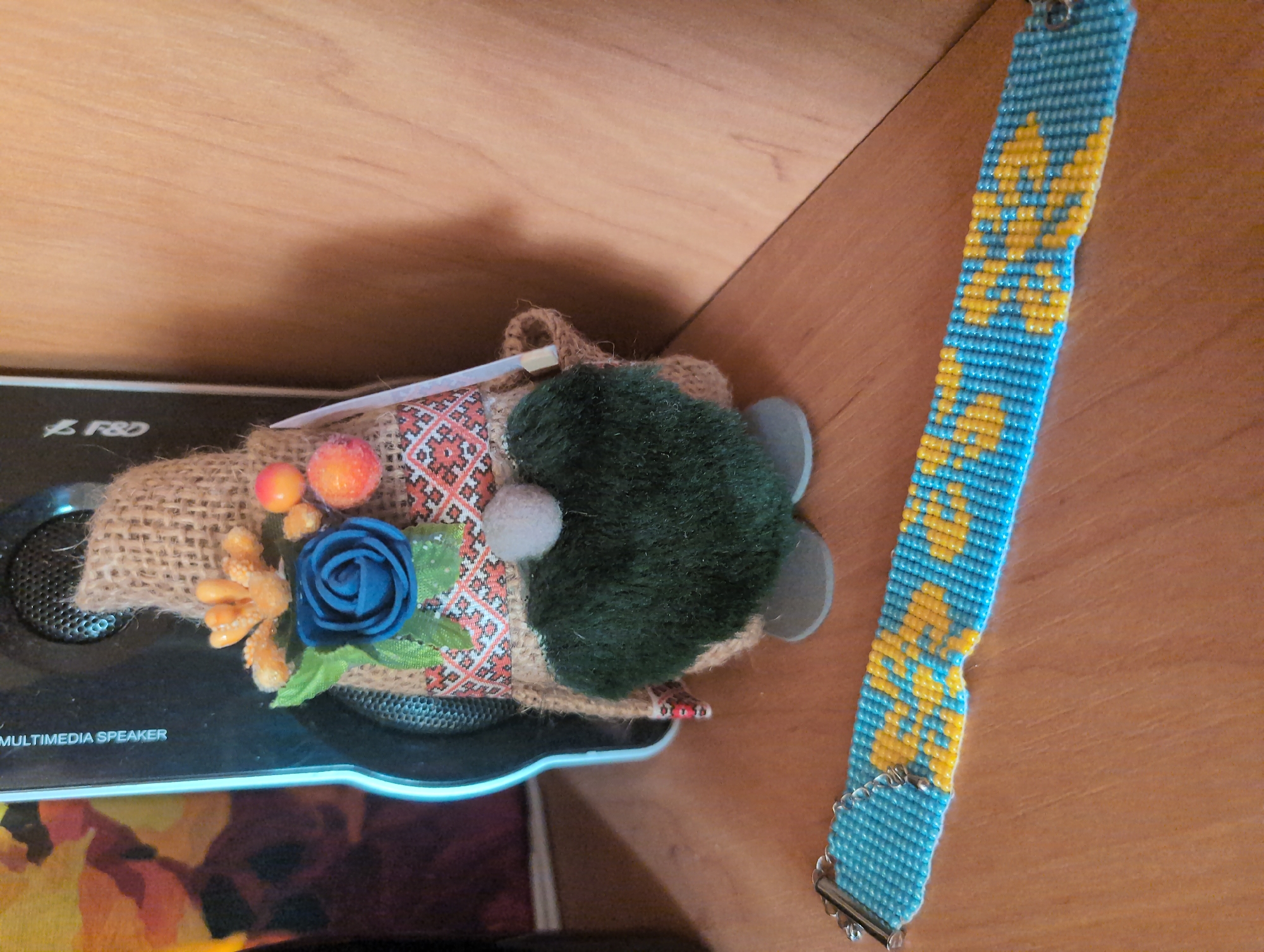 A beaded bracelet with a blue and yellow pattern lies on a wooden surface. Behind it, a gnome-like figurine is propped against a black speaker. The figurine has a burlap body, a dark green fuzzy beard, and is adorned with a blue flower and orange berries. The speaker has the words "MULTIMEDIA SPEAKER" printed vertically on its side. The background appears to be a wooden wall. This scene is located in Kryvyi Rih, Ukraine.