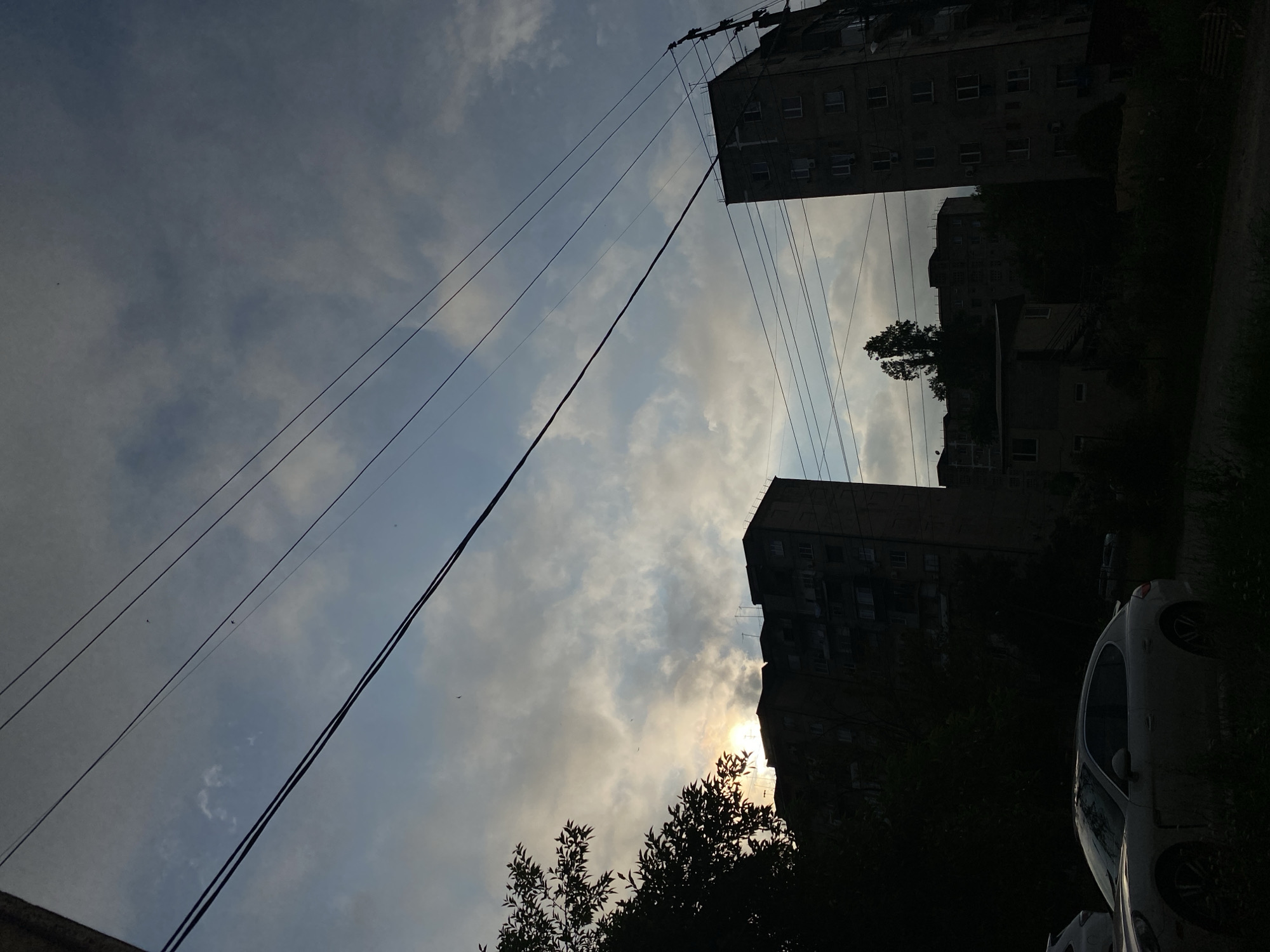 The image depicts a dimly lit outdoor scene, likely in a residential area of Unknown, Unknown, at dusk or dawn. In the foreground, a light-colored sedan is parked on a dirt or gravel surface, partially obscured by foliage. Behind the car, several multi-story apartment buildings are visible, their facades dark and indistinct. The sky above is a mixture of dark and lighter clouds, with a faint glow suggesting the presence of the sun behind them. Multiple power lines crisscross the frame, adding to the urban setting. The overall impression is one of a quiet, somewhat somber urban environment.