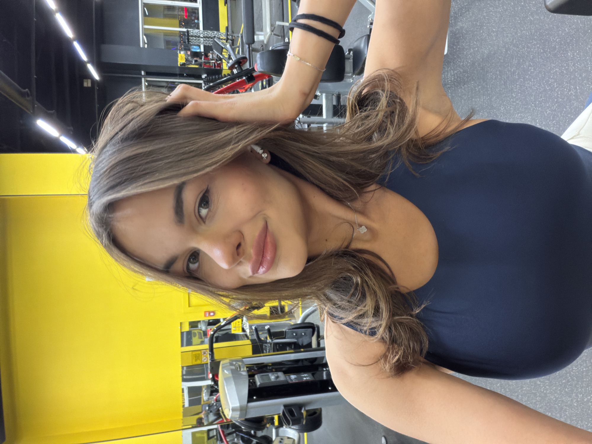 The image appears to be a selfie taken in a gym, likely located in Krasnoyarsk, Russia. A woman with long brown hair, wearing a dark blue athletic top, is facing the camera. Her right hand is raised, touching her hair. Behind her is typical gym equipment, including weight machines. A yellow wall is visible on the left. The floor is gray. She's wearing a necklace and has a couple of bands on her wrist.
